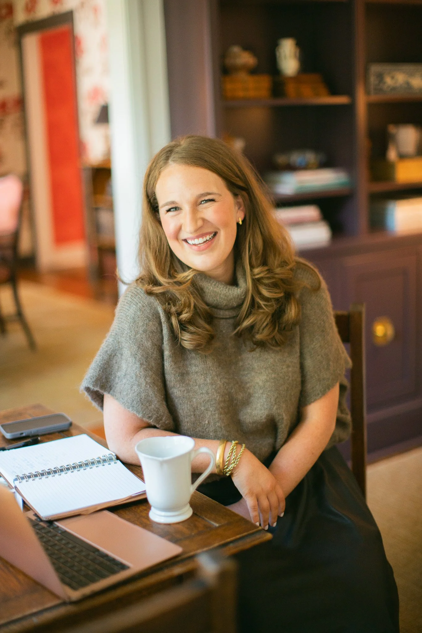 A woman with long brown hair, smiling and sitting at a wooden table in a cozy indoor setting, with a mug, notebook, and phone on the table, and a cabinet with books and decorative objects in the background.