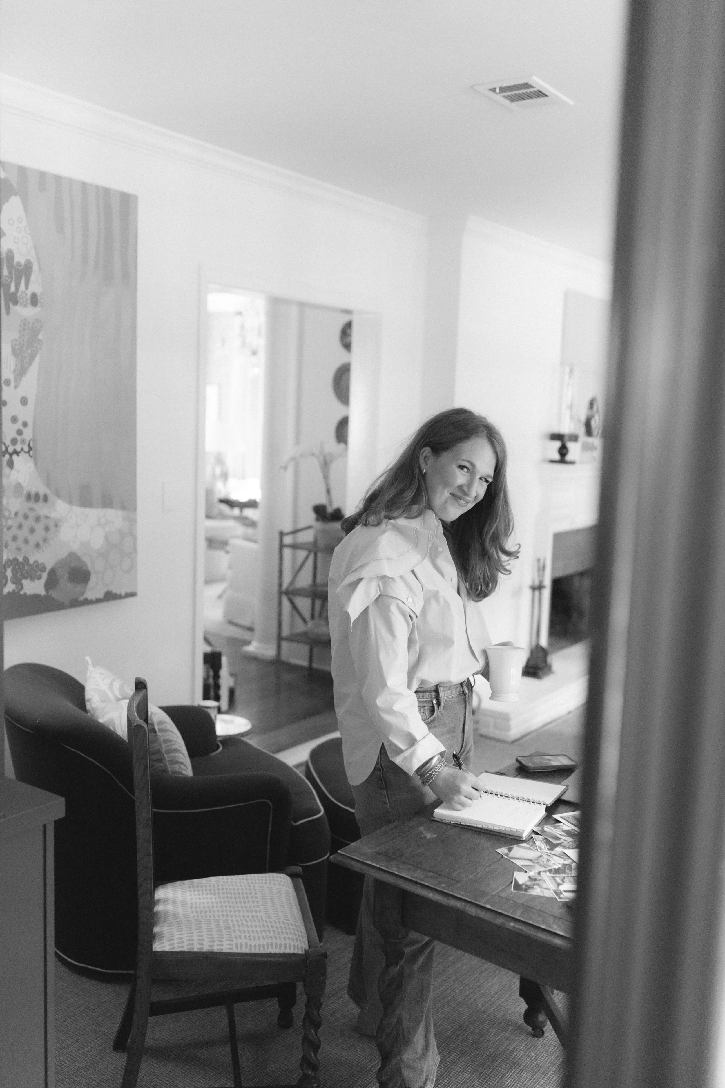 A woman with long hair, smiling, standing in a living room near a dining table, writing in a notebook and holding a cup.