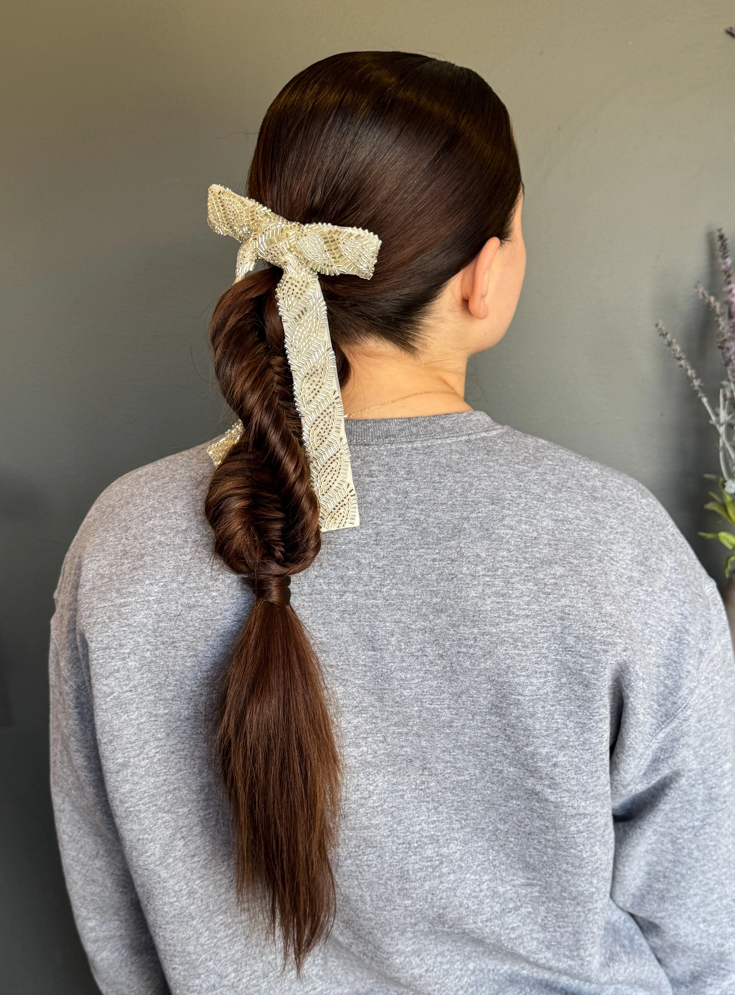 Back view of a woman with long brown hair in a braid, accessorized with a cream-colored sequin bow hair ribbon, wearing a gray sweatshirt, standing in front of a gray wall with some flowers in the background.