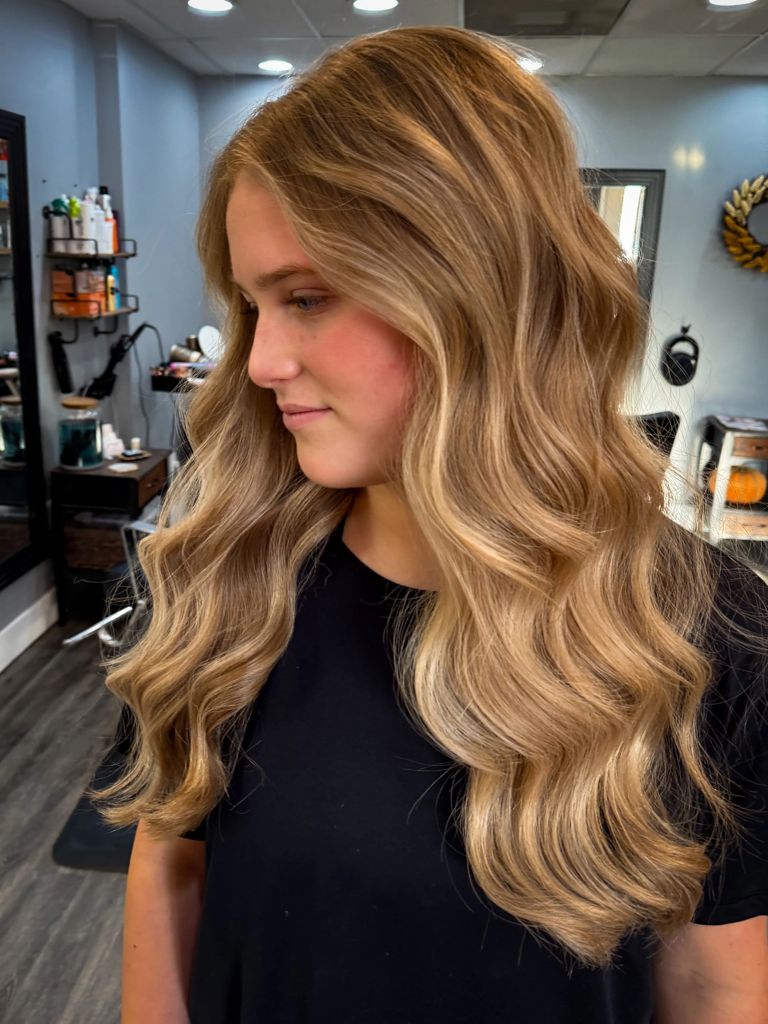 Side profile of a woman with long, wavy blonde hair, wearing a black top, inside a hair salon.