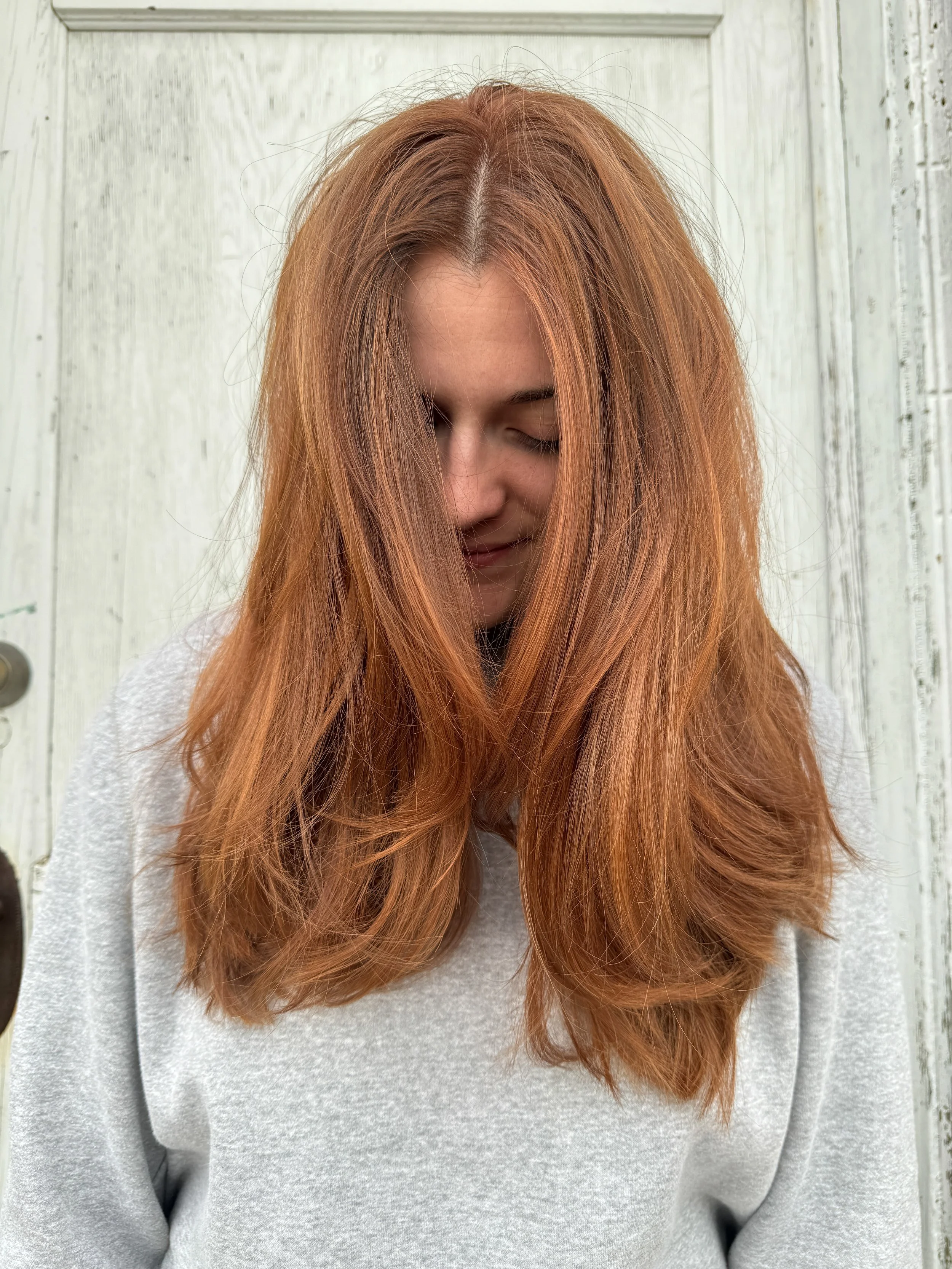 A woman with long, wavy, strawberry blonde hair wearing a gray sweatshirt, standing against a light-colored wooden background. She has her eyes closed and wears a slight smile.