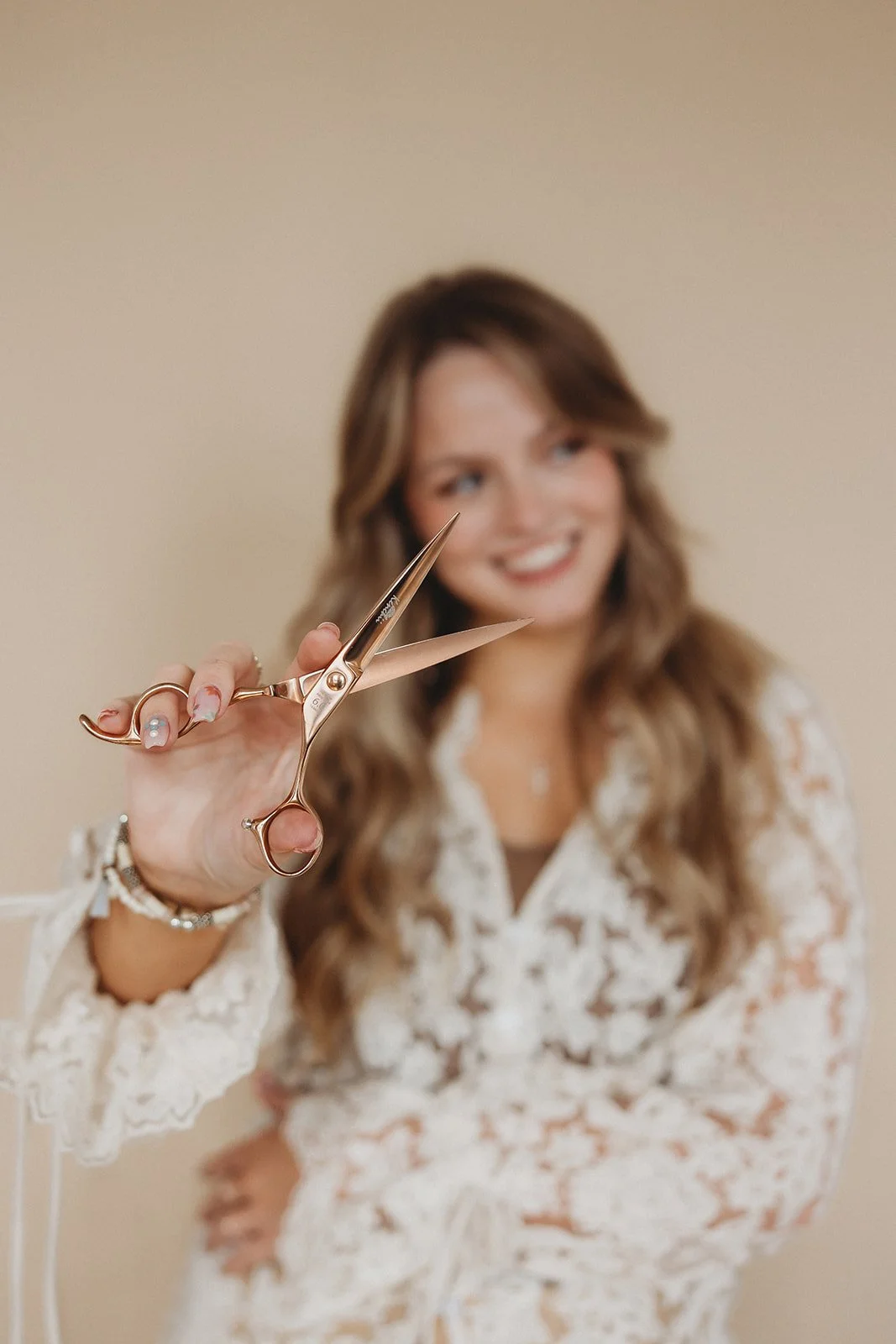 Woman with long wavy hair holding a pair of scissors towards the camera, smiling, wearing a lace blouse, against a beige background.