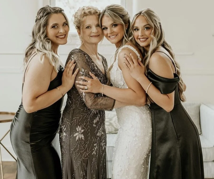 A mother and 3 daughters, one of which is a bride, with their hair styled, smiling at the camera