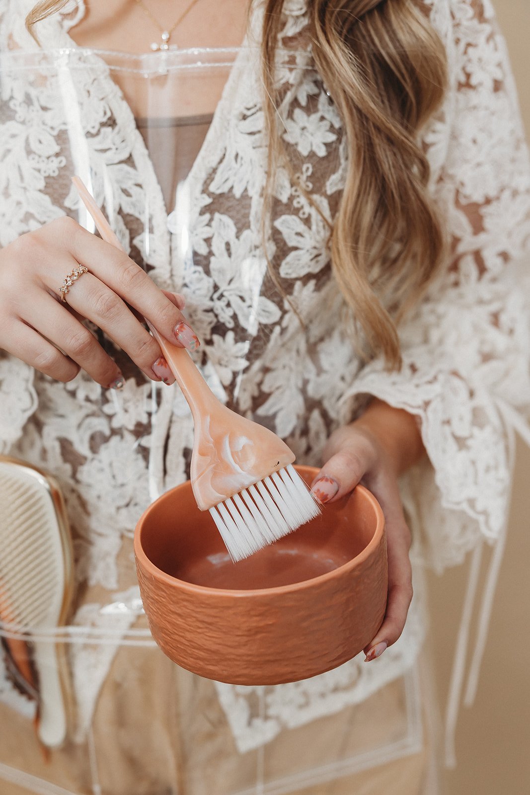 A woman wearing a lace dress is holding a small terracotta bowl and mixing hair color with a color brush.