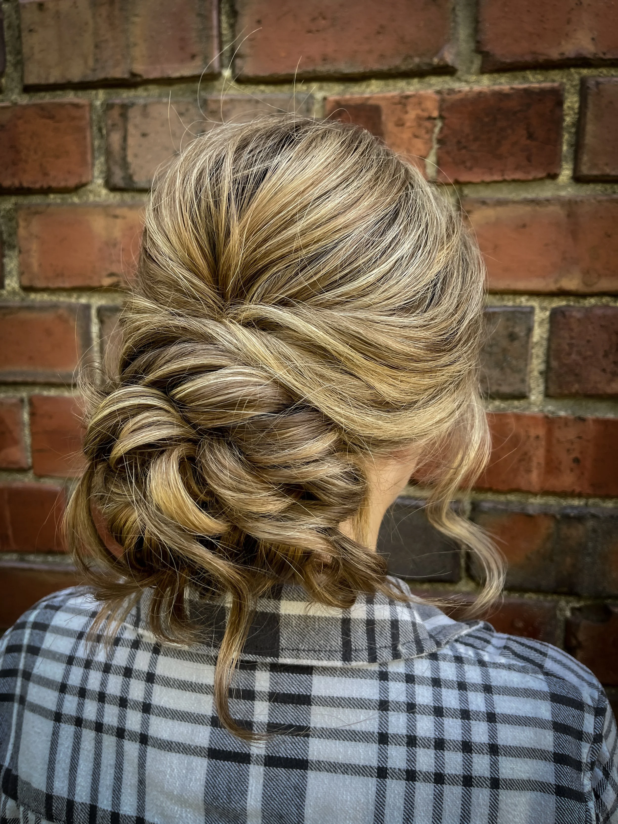 Back of a woman's head with her hair styled in loose, voluminous curls, wearing a plaid shirt, against a brick wall background.