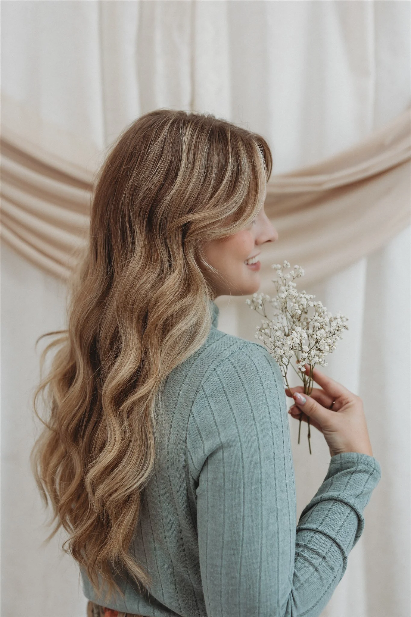 A woman with long, wavy blonde hair holding a small bouquet of white baby's breath flowers, wearing a light blue, ribbed, long-sleeve shirt, standing in front of a beige draped curtain.