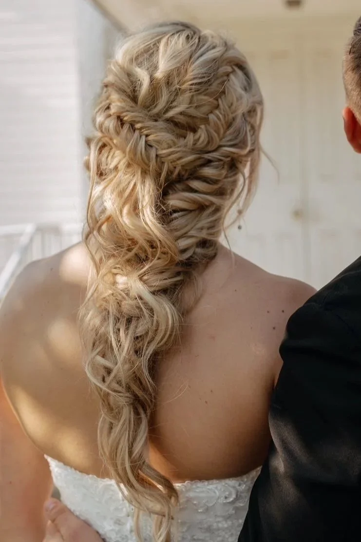 Back view of a bride with long, blonde, braided and curled hair, wearing a strapless white dress, beside a groom in a black suit.