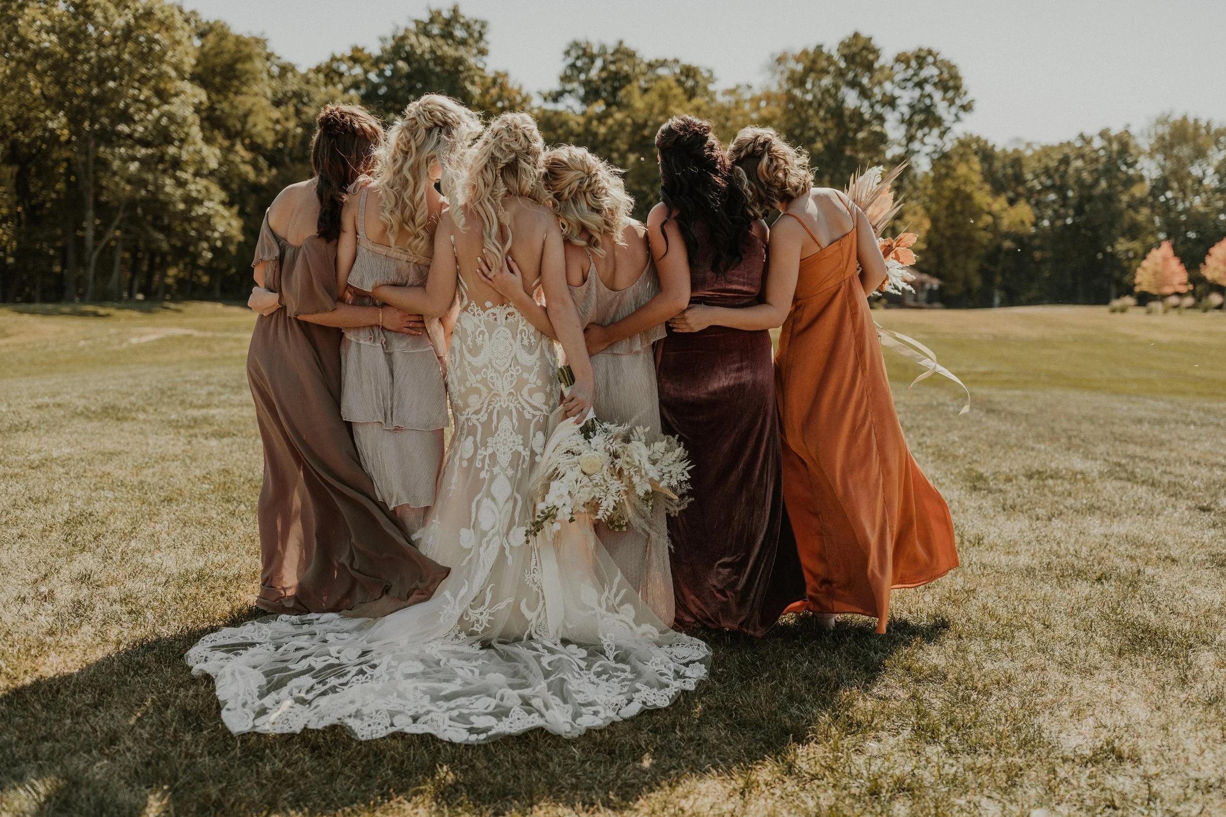 Group of women, including a bride in a white lace wedding dress holding a bouquet, standing closely together outdoors on a grassy field with trees in the background.