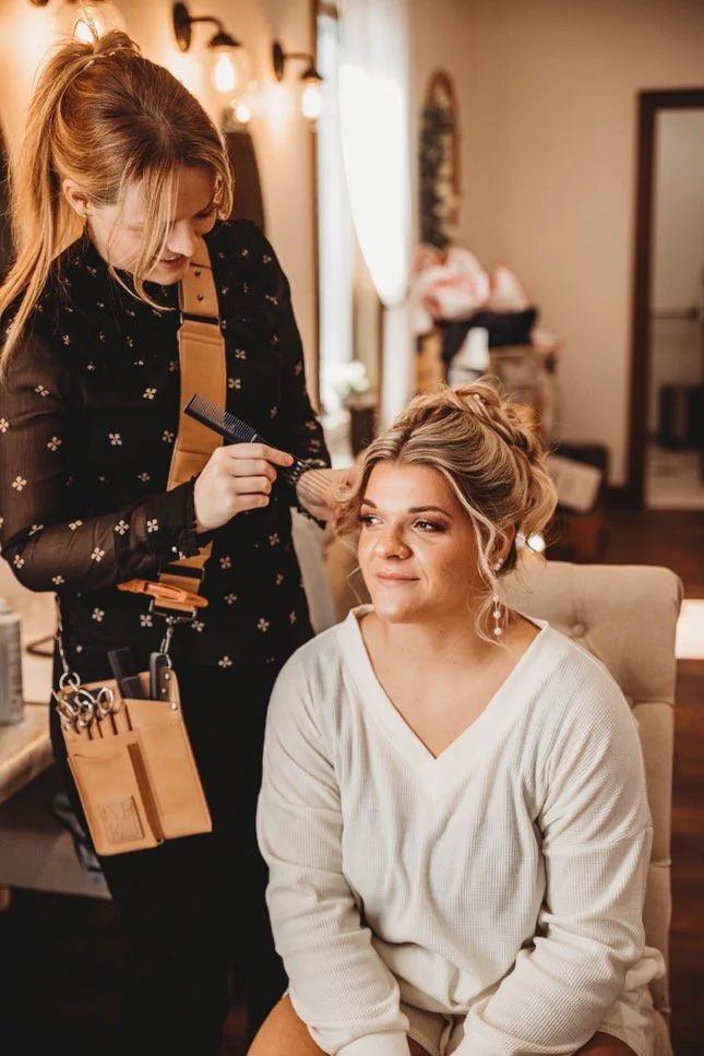 A woman is sitting in a chair with her hair styled by a hairstylist who is combing her hair, in a cozy room with warm lighting.