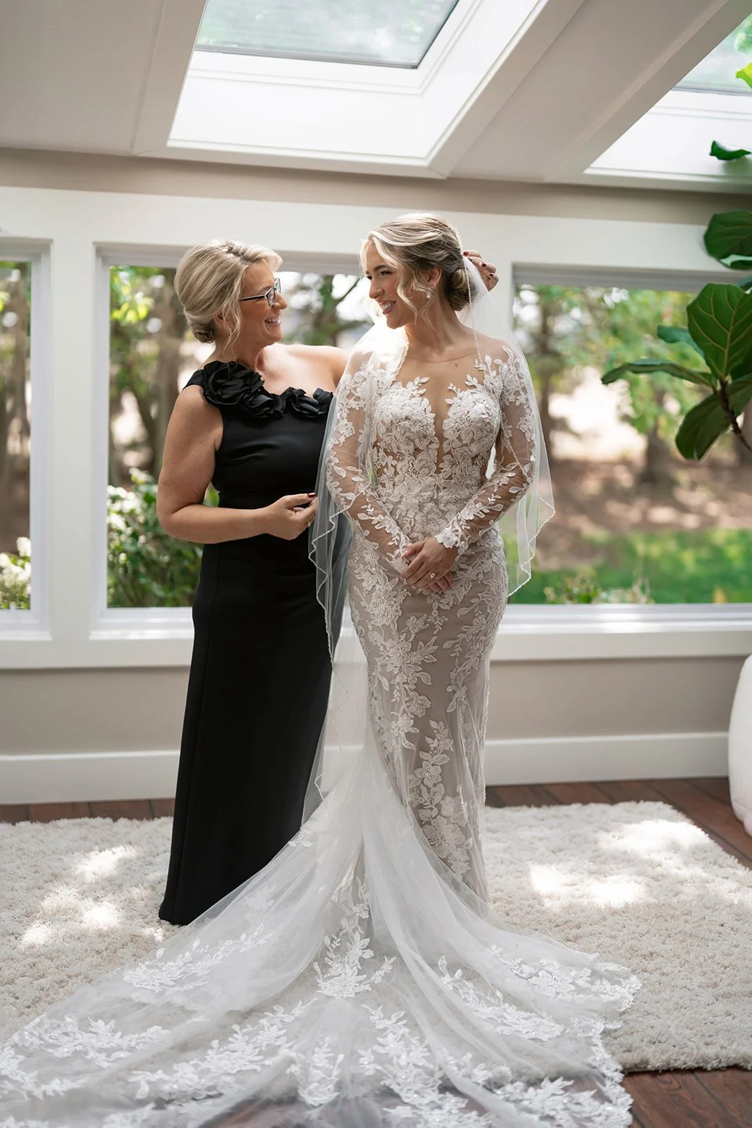 A bride in a lace wedding gown with long sleeves stands next to the mother of the bride in a black dress, inside a room with large windows and skylights, greenery outside, and a white rug on the floor.