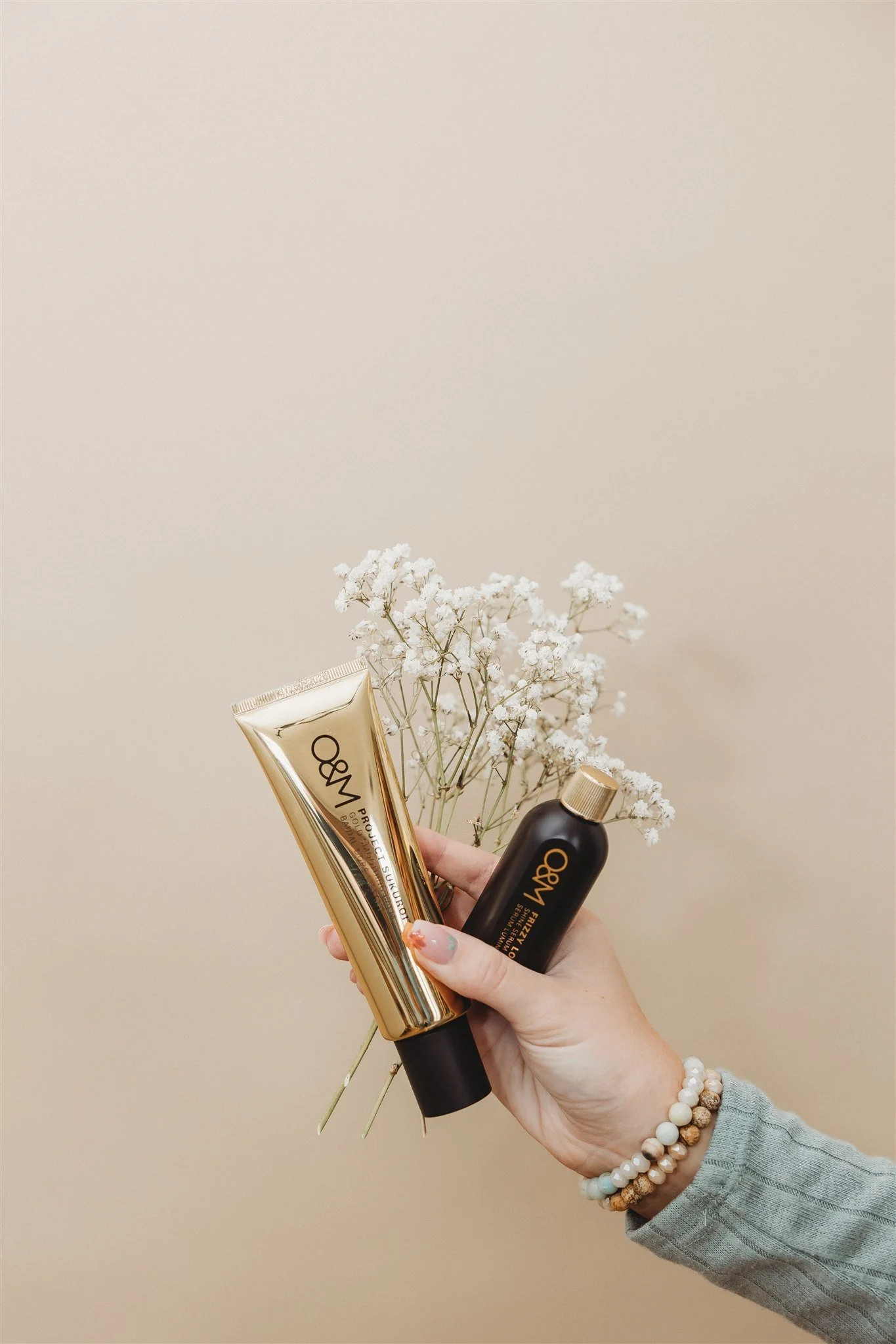 Hand holding a gold tube and a black bottle of O&M hair products in front of white baby's breath flowers and a beige background.