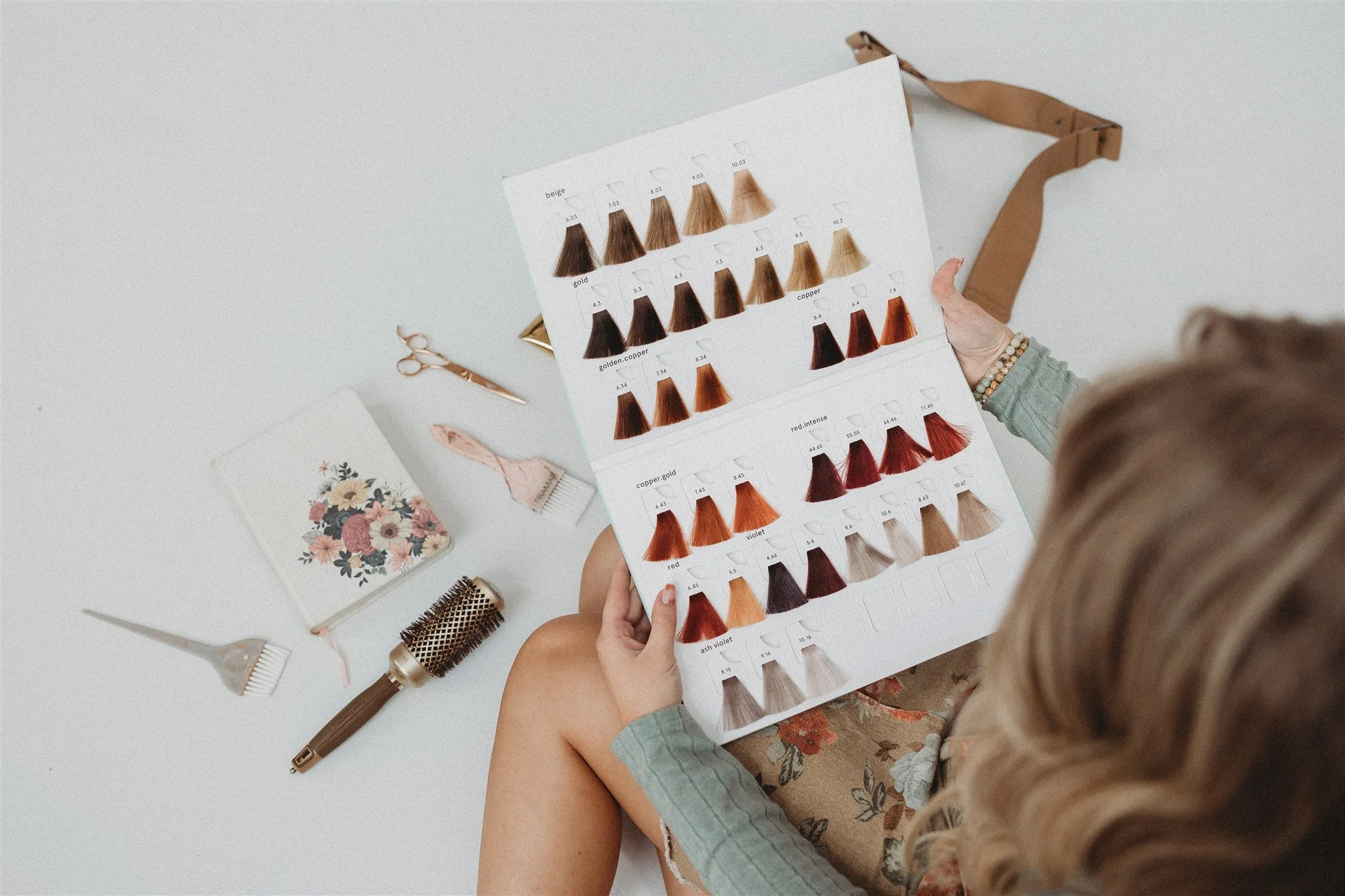 A woman looking at a hair color chart with various shades of hair dye, surrounded by hair styling tools and accessories on a white surface.