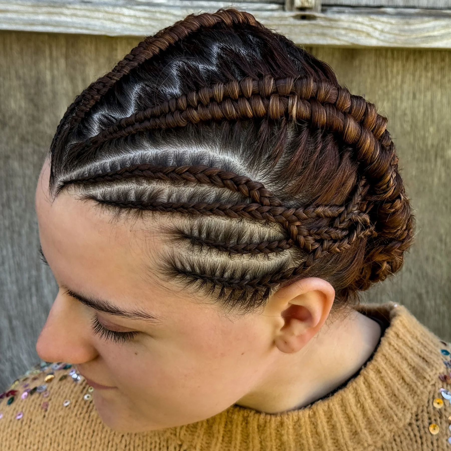 A woman with intricately braided and styled hair, featuring multiple cornrows and twisted sections, styled in an updo, wearing a beige sweater with sequins.