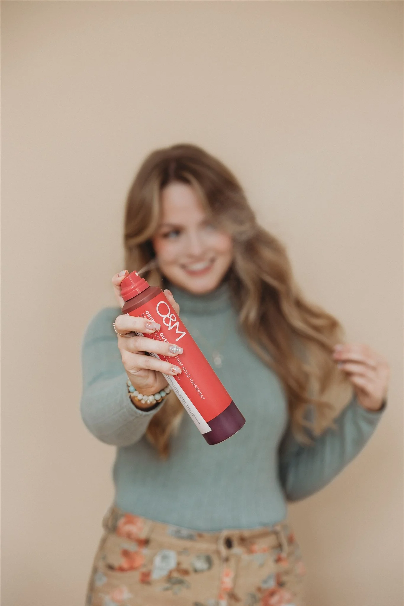 A woman with long, wavy hair holding a red can of hair spray, smiling, with a beige background.