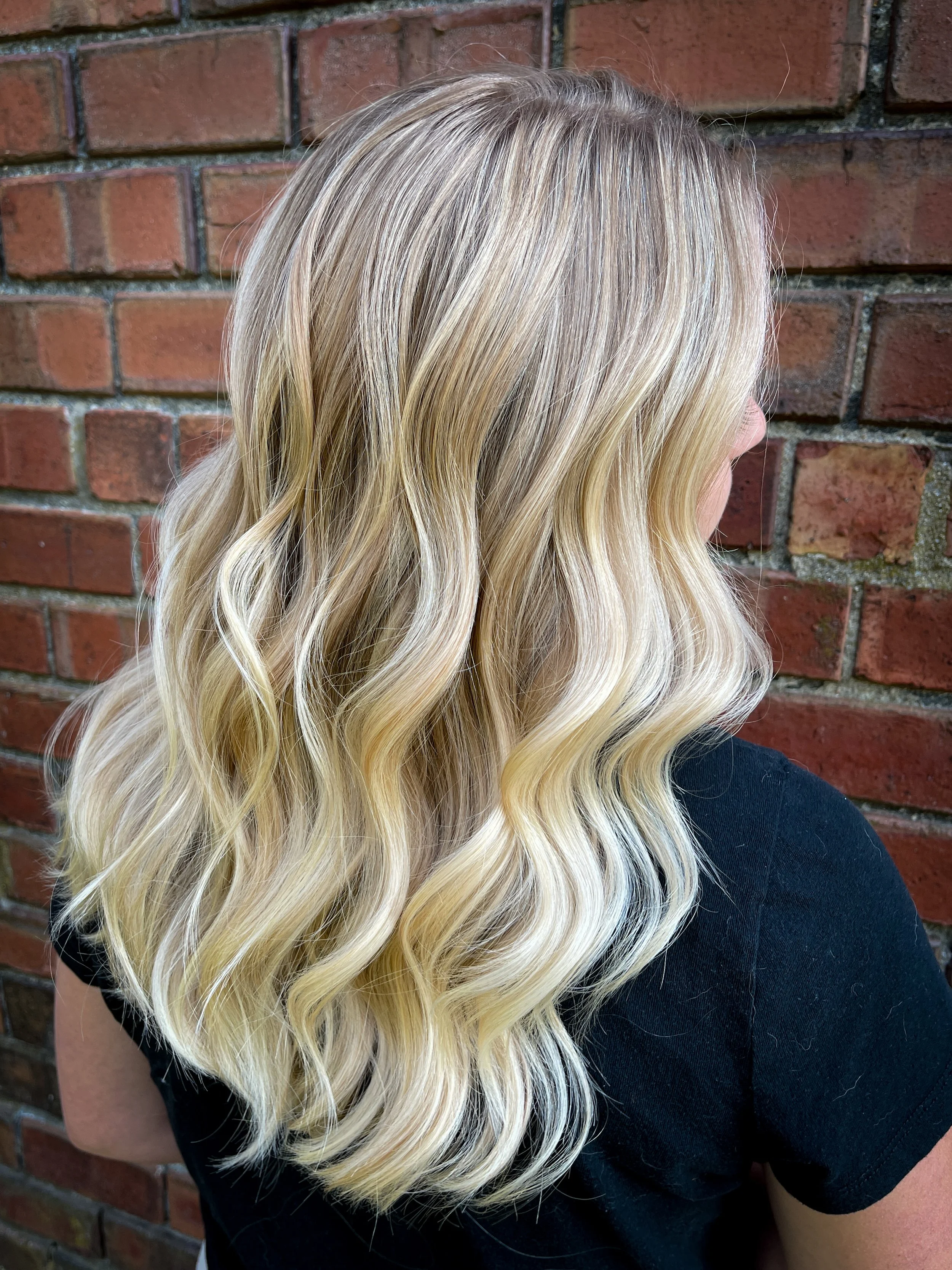 A woman with long, wavy blonde hair standing against a brick wall.