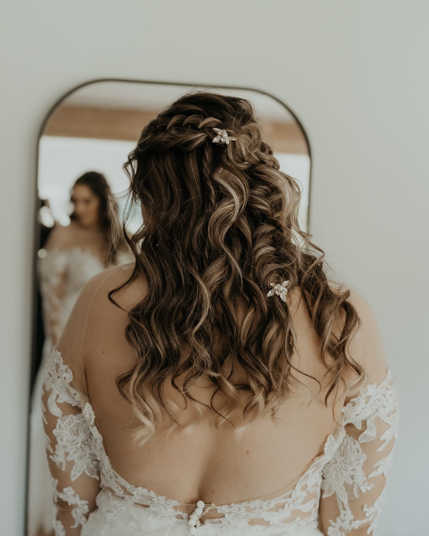 Brunette woman with long, curly hair decorated with butterfly clips, wearing a lace wedding dress, looking into a mirror, with her reflection visible.