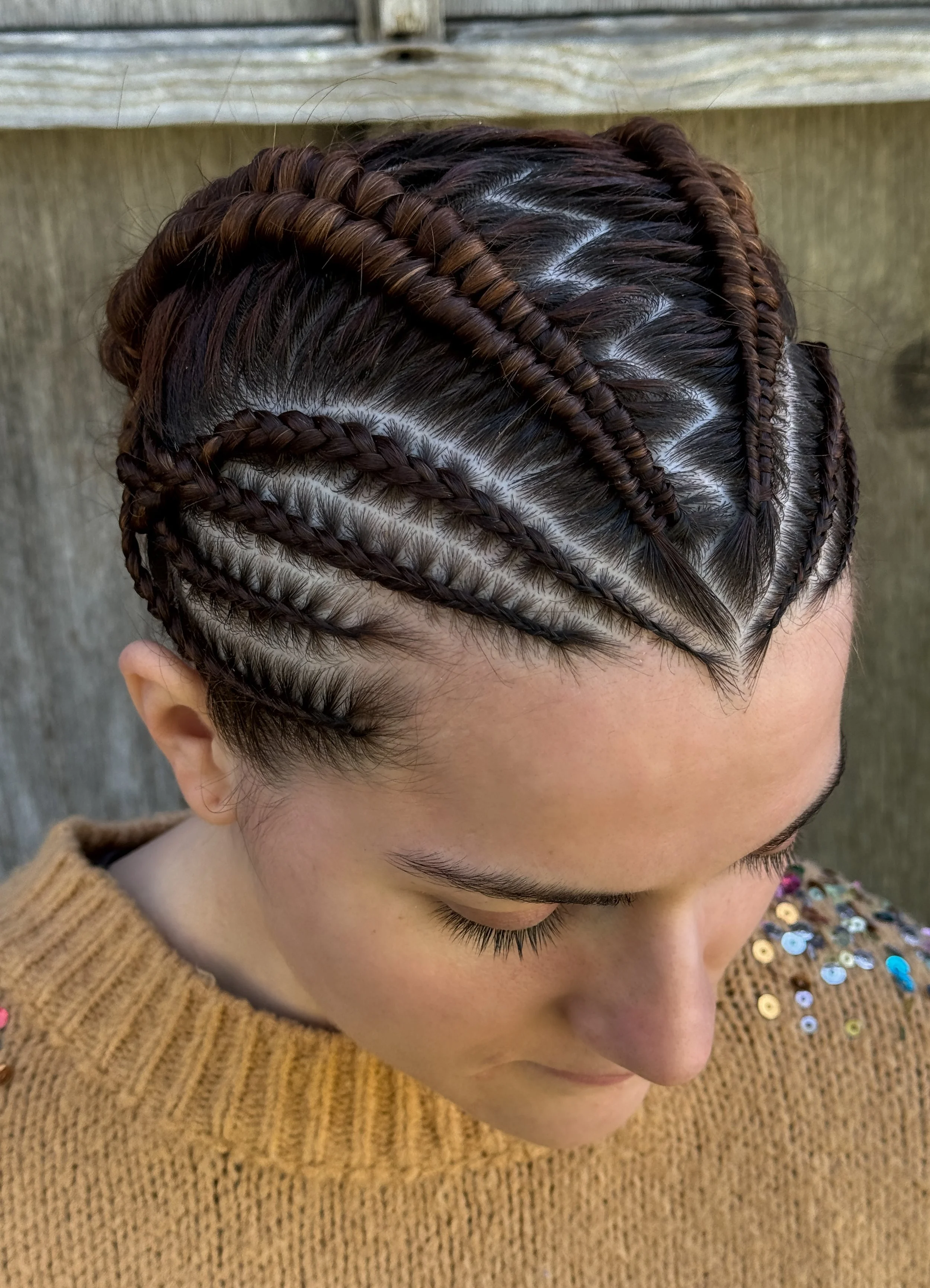 Close-up of a woman with intricately braided hairstyle and dark brown hair, wearing a brown sweater with colorful sequins on the shoulders, standing in front of a wooden fence.