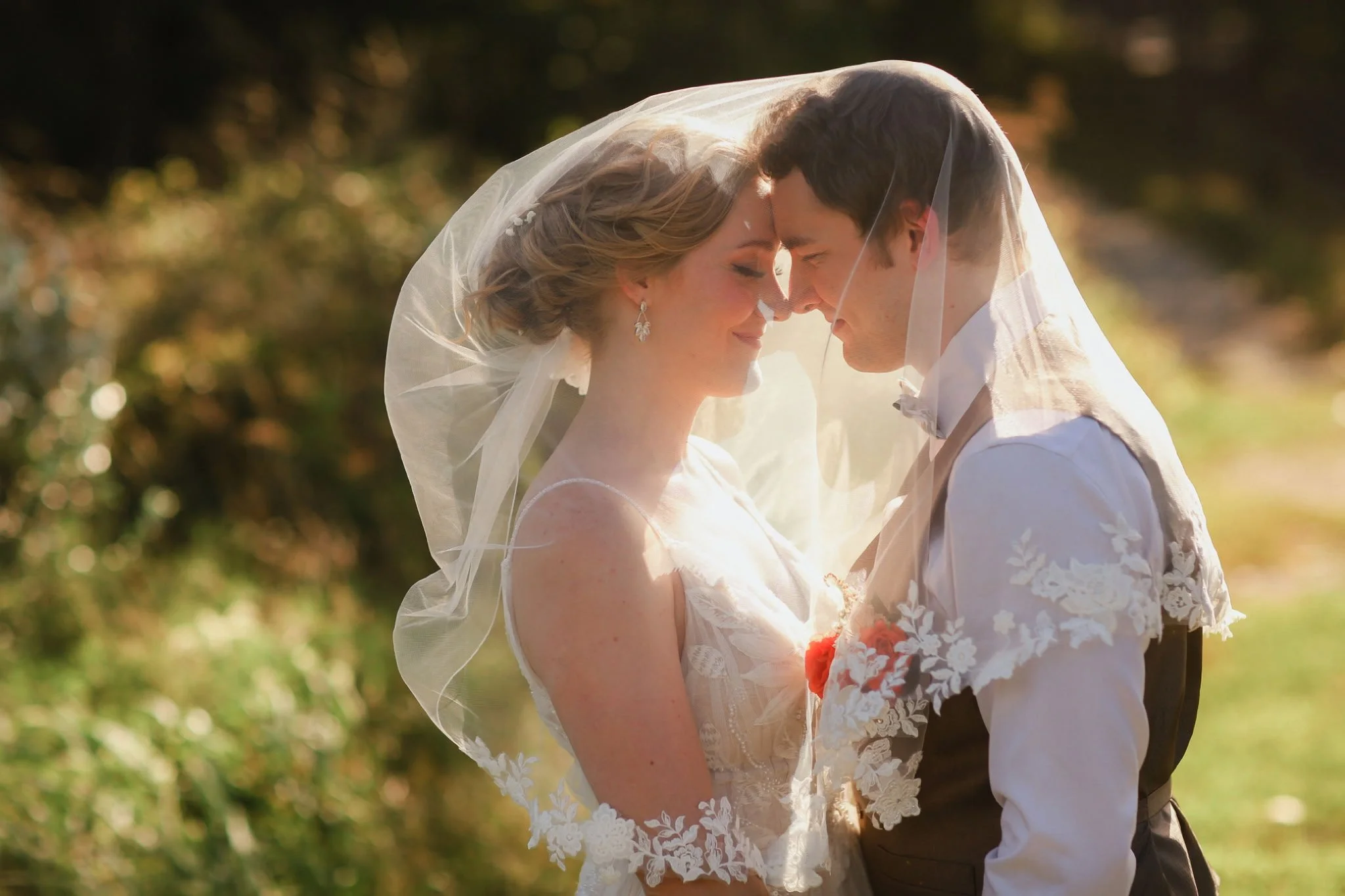 A bride and groom standing close with their foreheads touching, smiling, under a veil outdoors during sunset.