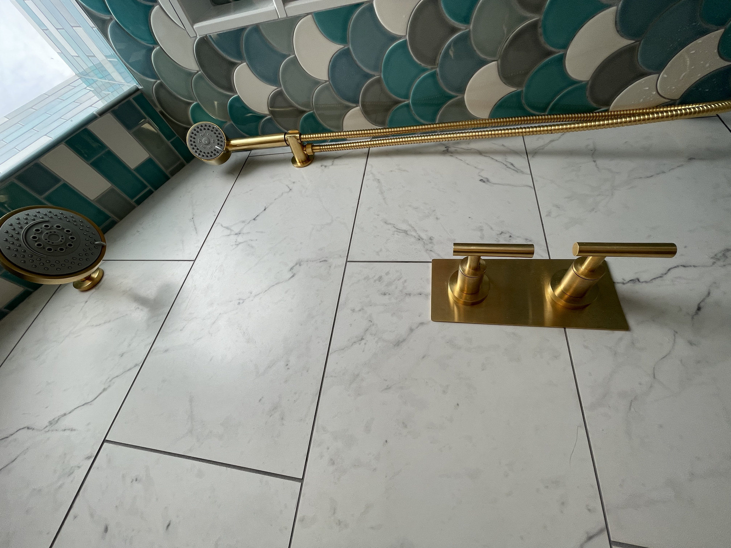 Bathroom shower with gold fixtures, including a dual-handle faucet and a handheld showerhead, with a decorative tiled wall and white marble floor tiles.