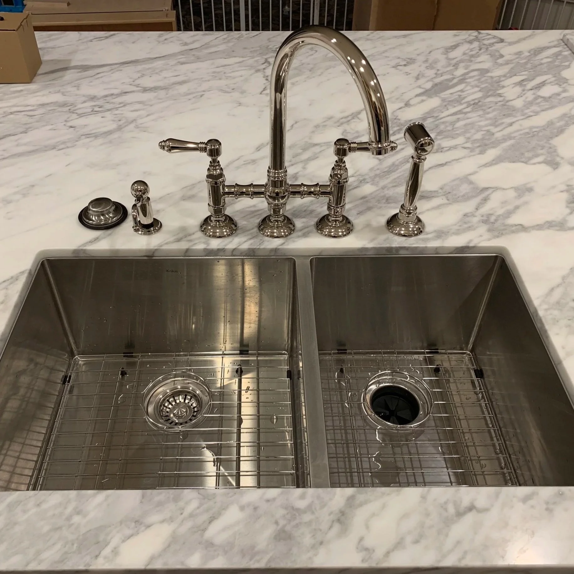 A kitchen sink with two metal basin and a marble countertop. The faucet has multiple handles and a spray nozzle.