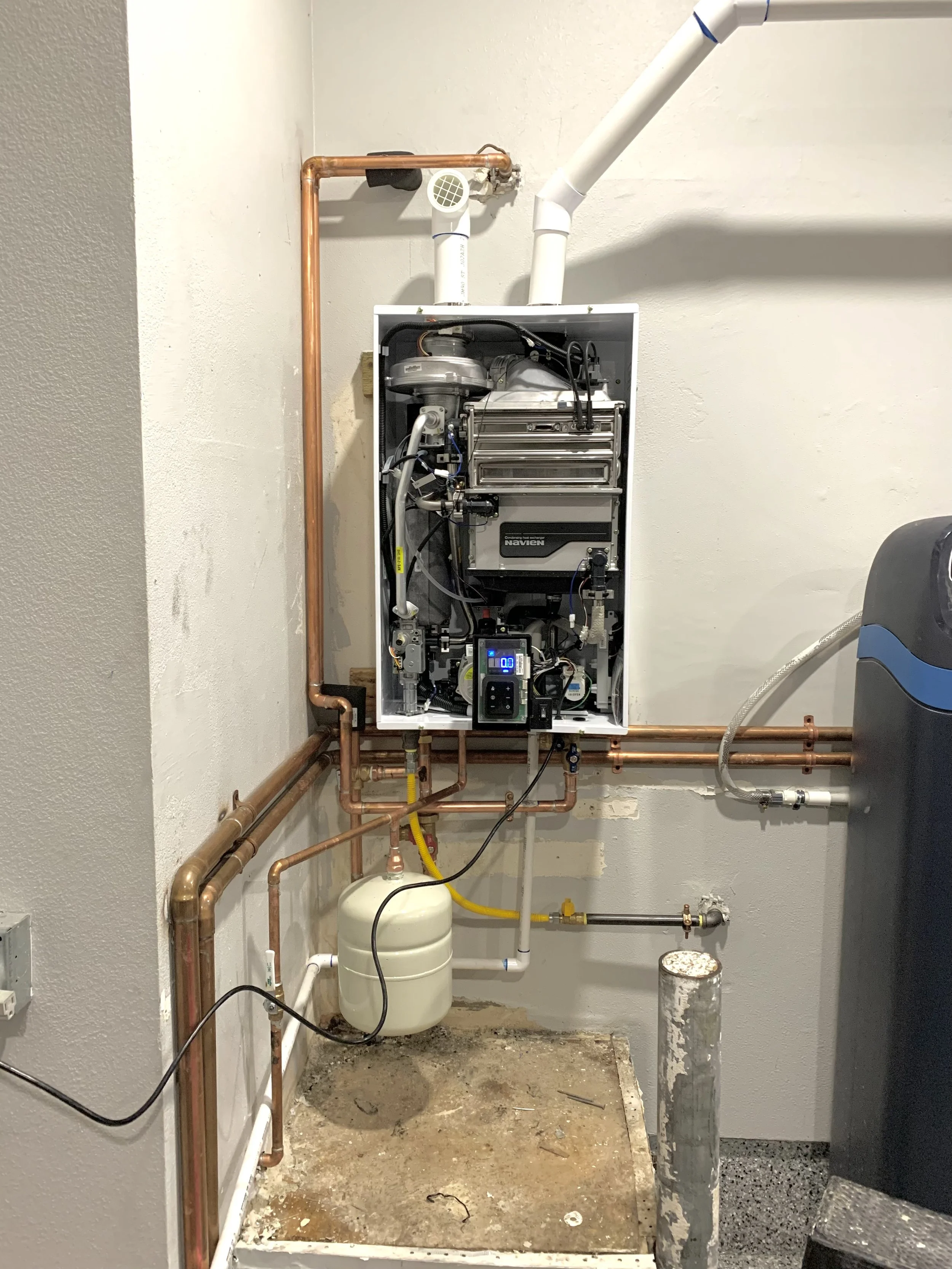 Interior view of a boiler system with exposed copper pipes, an open control panel, and various connected water and gas lines.
