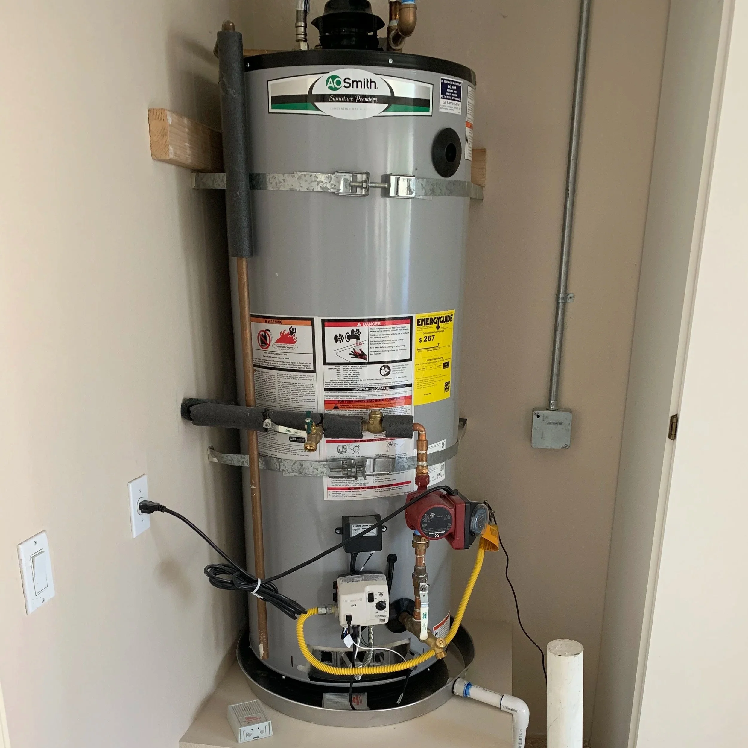 A new, gray AO Smith water heater installed in a utility closet with visible pipes, wires, and a yellow flexible gas line.