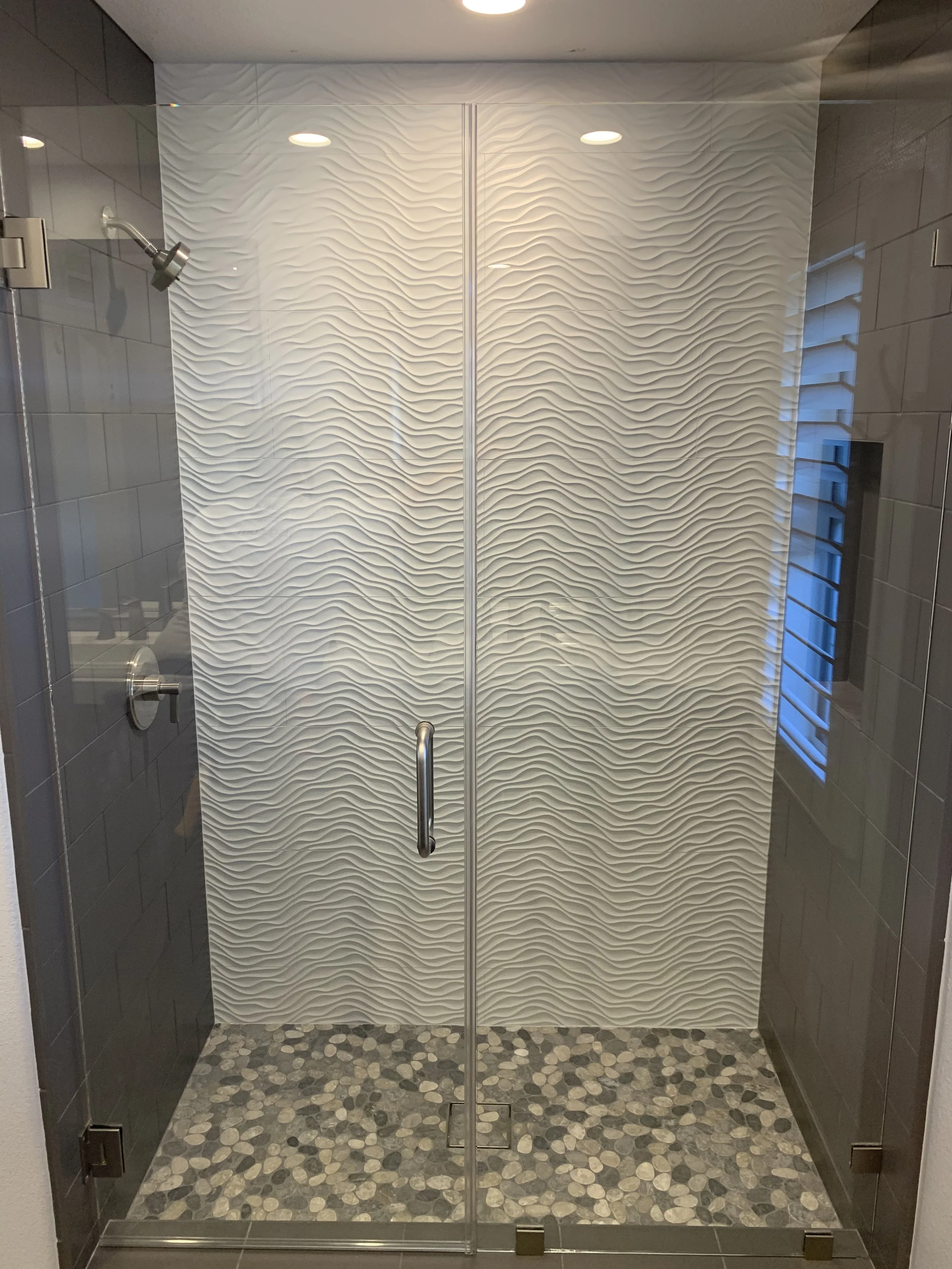 Shower stall with white wavy porcelain tile, river rock floor, and frameless glass doors