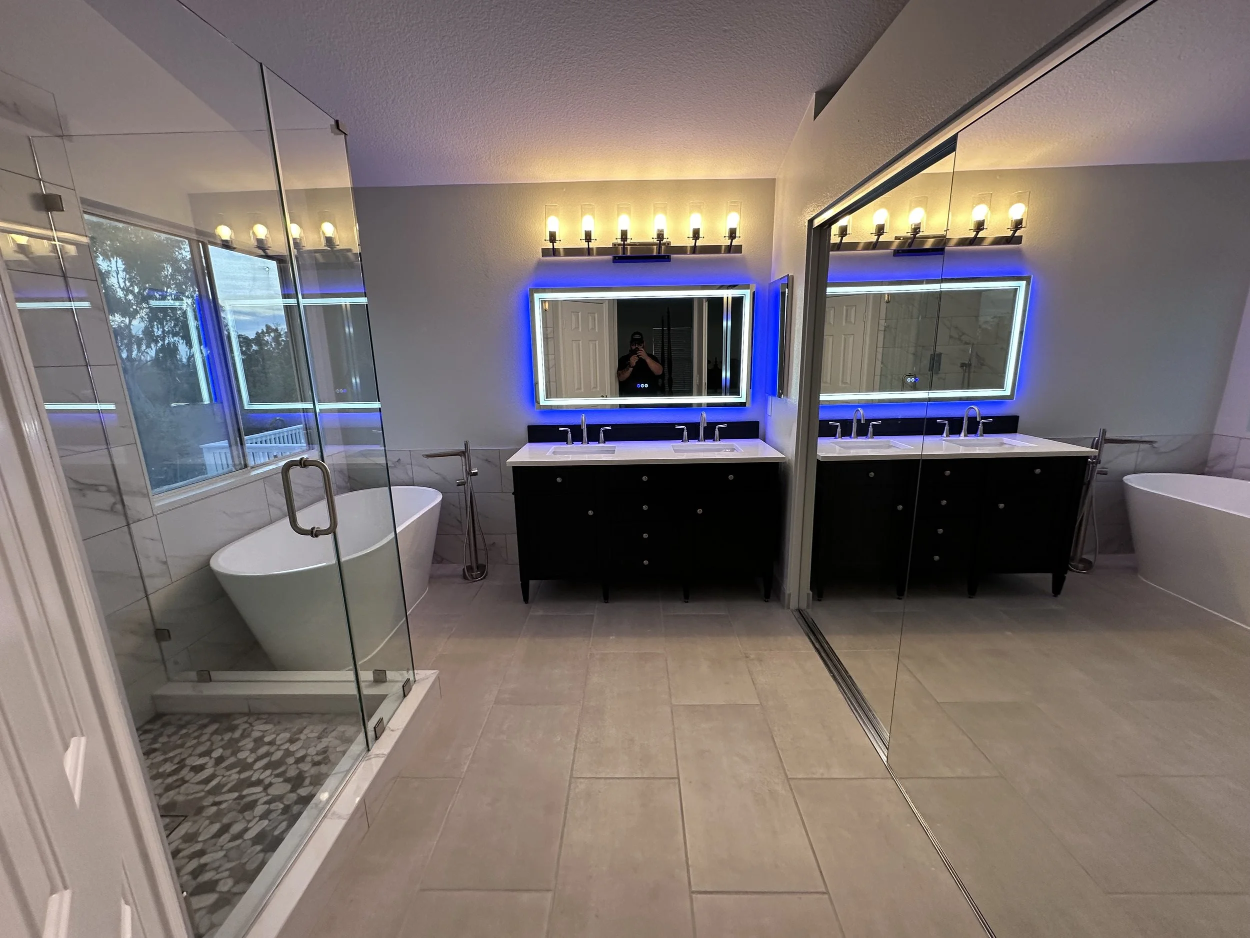 A large bathroom with tile floor, shower, freestanding tub, double vanity with a backlit LED mirror