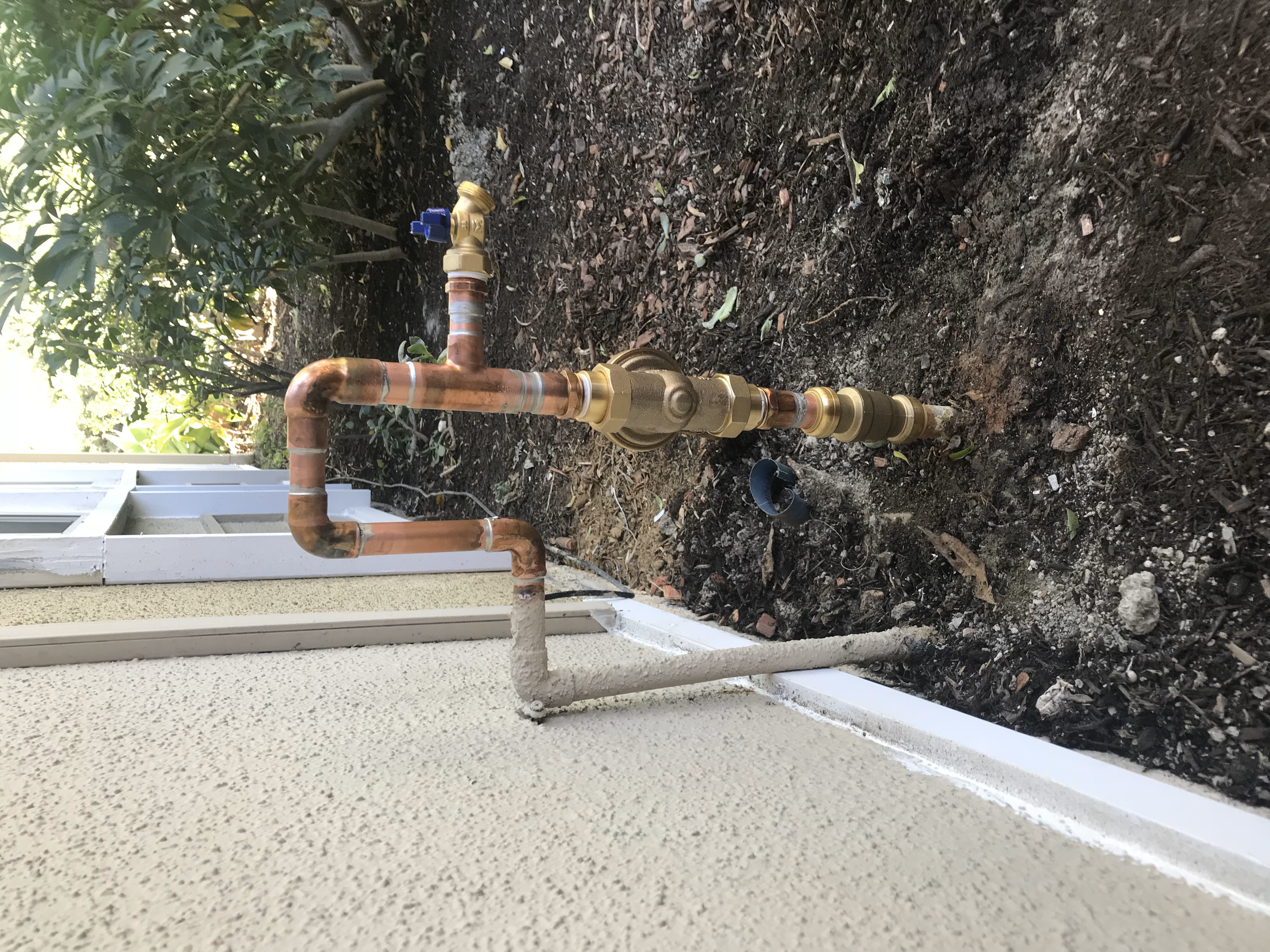 Copper plumbing pipes with brass fittings and a water valve installed next to a house exterior, with soil and plants in the background.