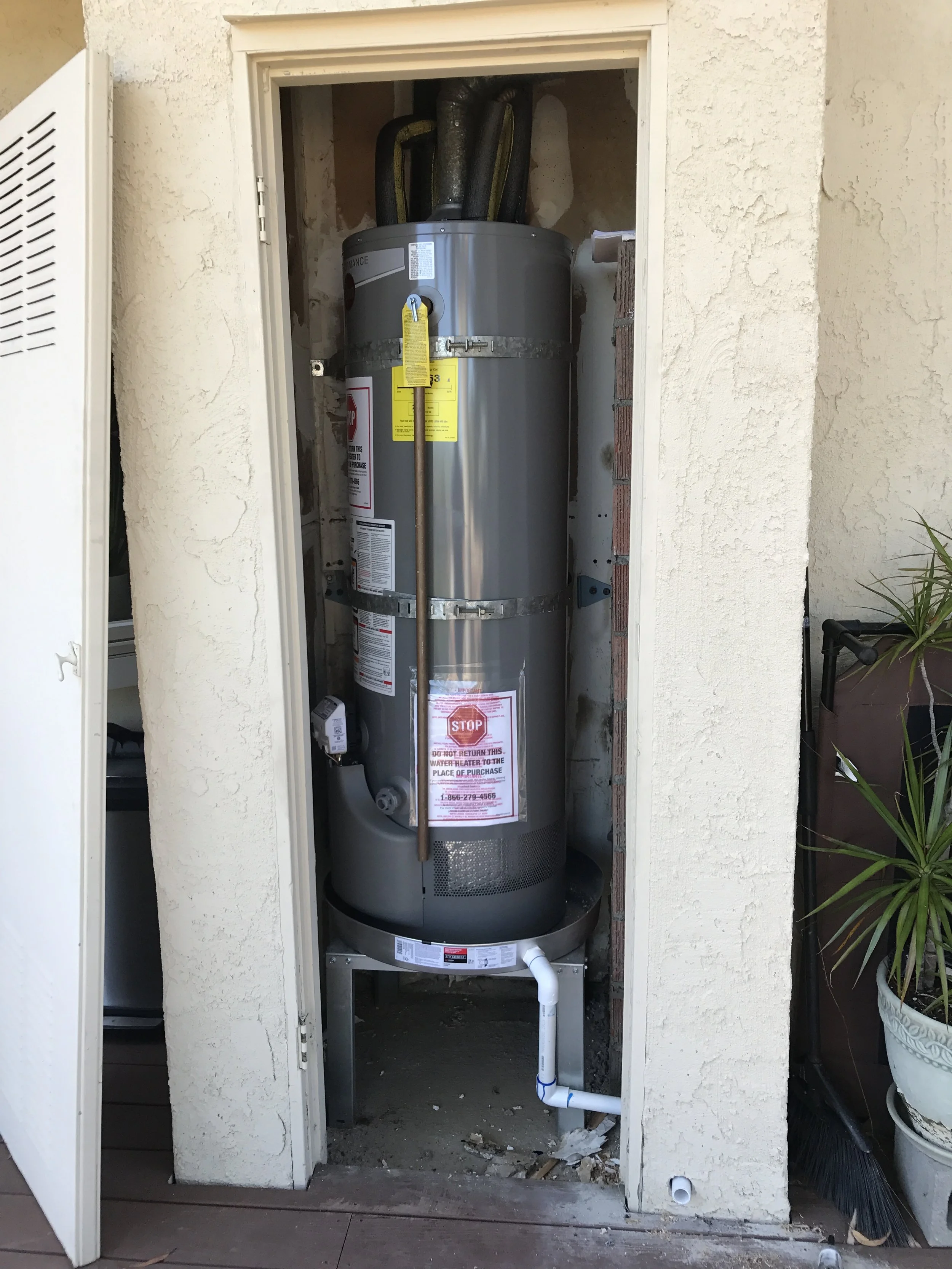 A water heater installed inside a small outdoor closet with an open door, showing pipes and warning labels.