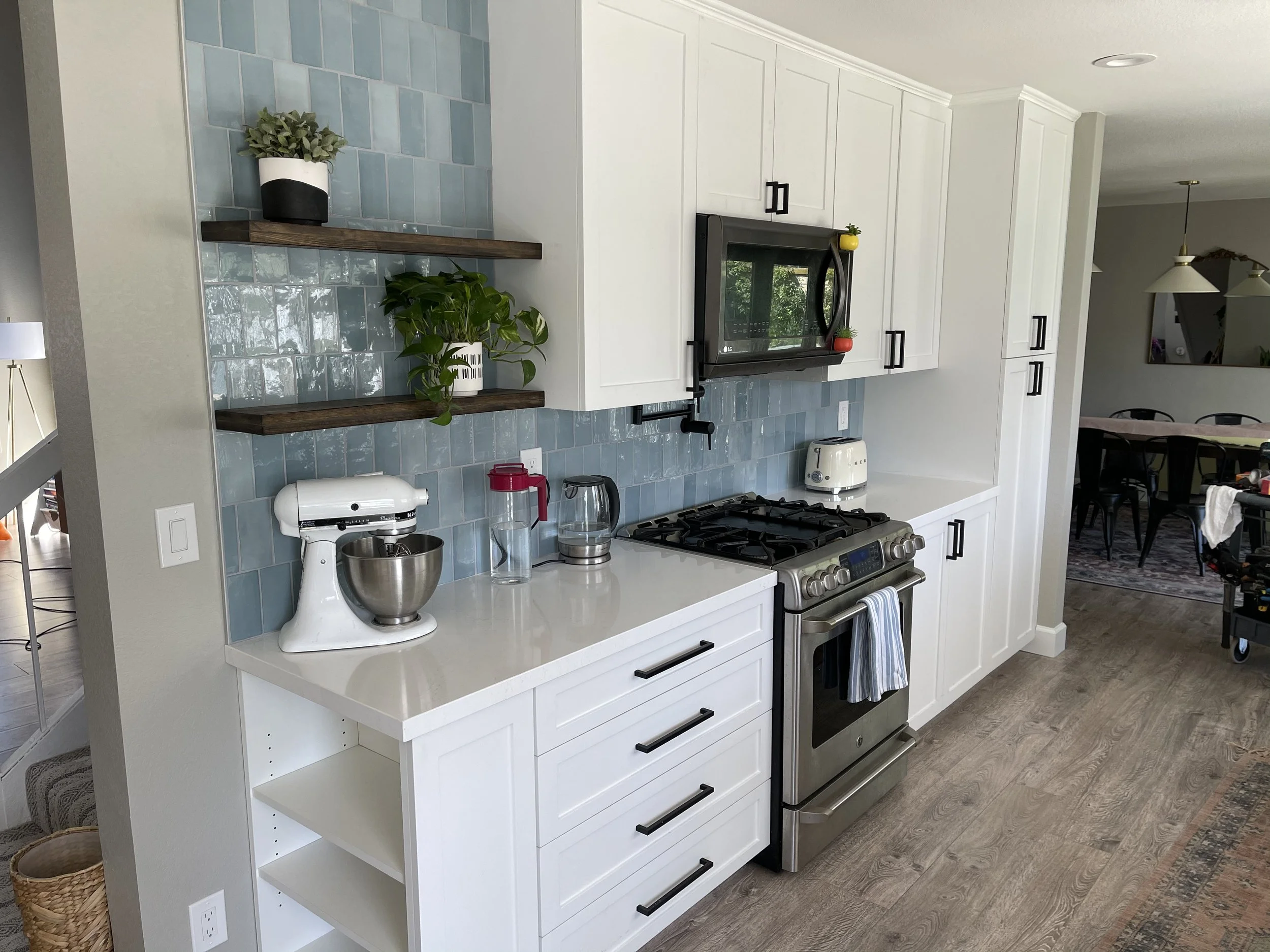Modern kitchen with white cabinets, light blue tiled backsplash, stainless steel stove and microwave, wooden open shelves with plants, small appliances on counter, and a dining area in the background.