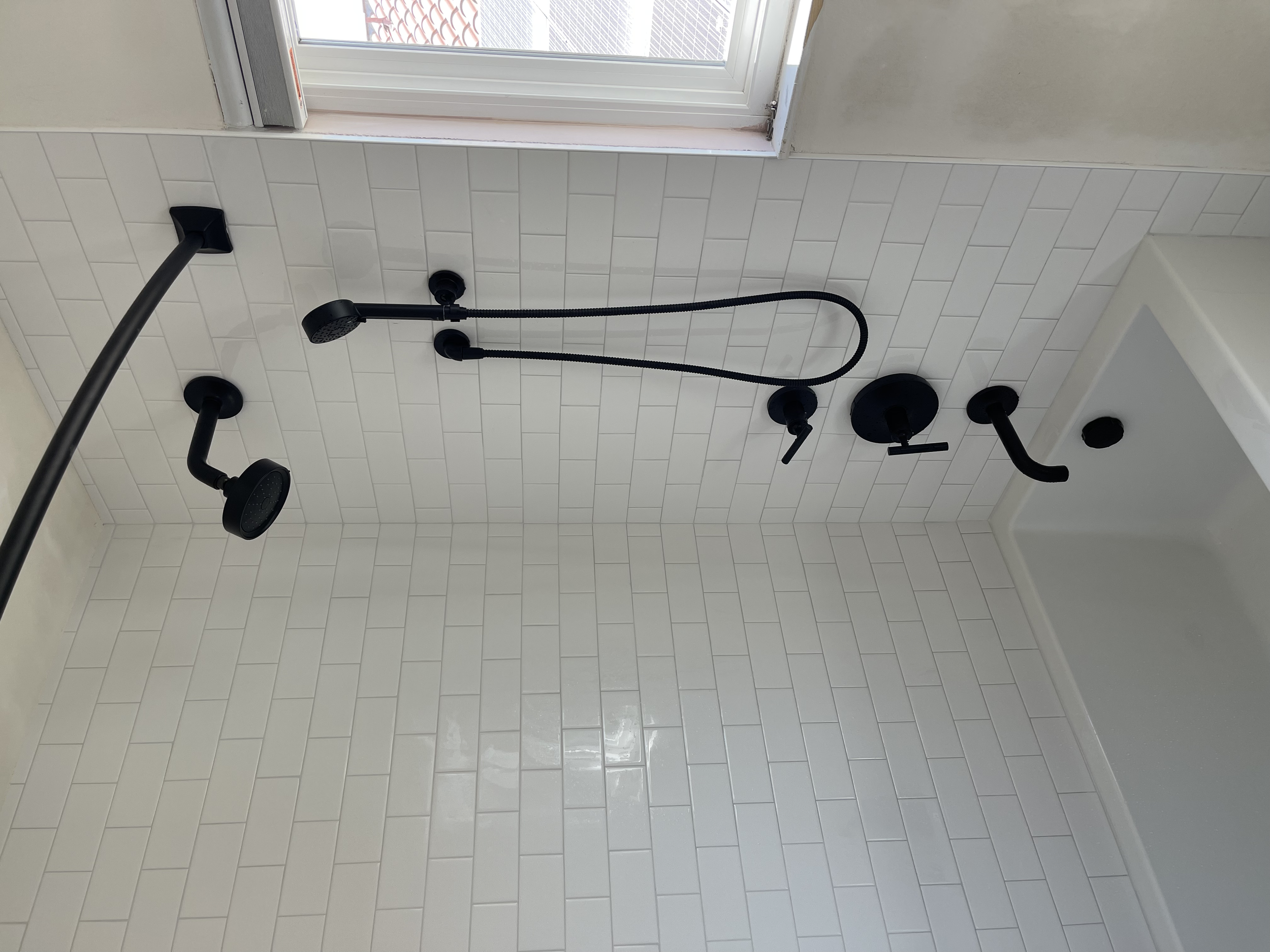 Black shower fixtures on a white tiled bathroom wall, including a handheld showerhead, a shower arm, a diverter lever, a laundry sink faucet, and a flexible hose, with a window at the top.