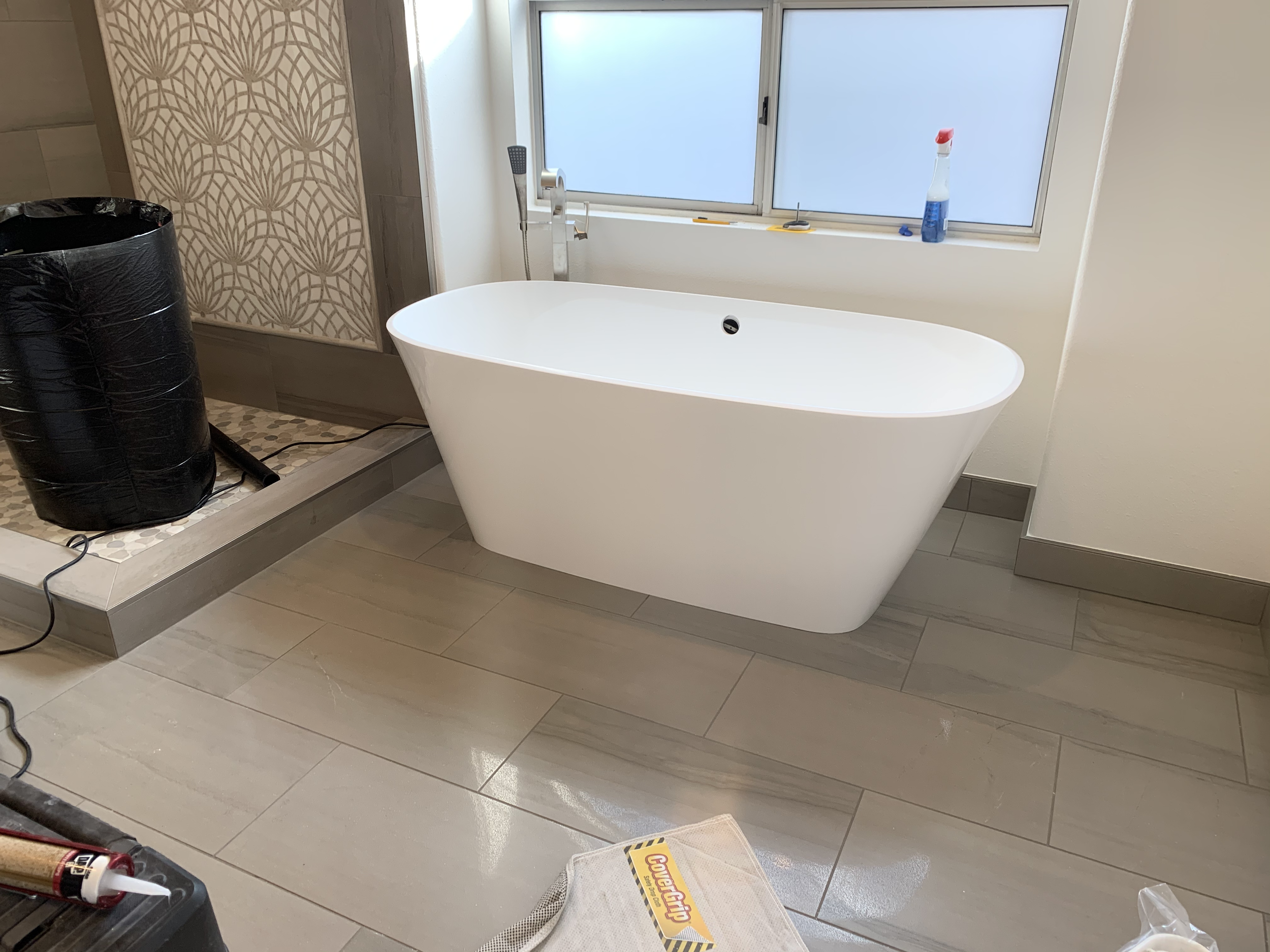Modern bathroom with white freestanding bathtub near a window, with cleaning supplies on the windowsill, a black trash can, and construction tools and materials nearby.