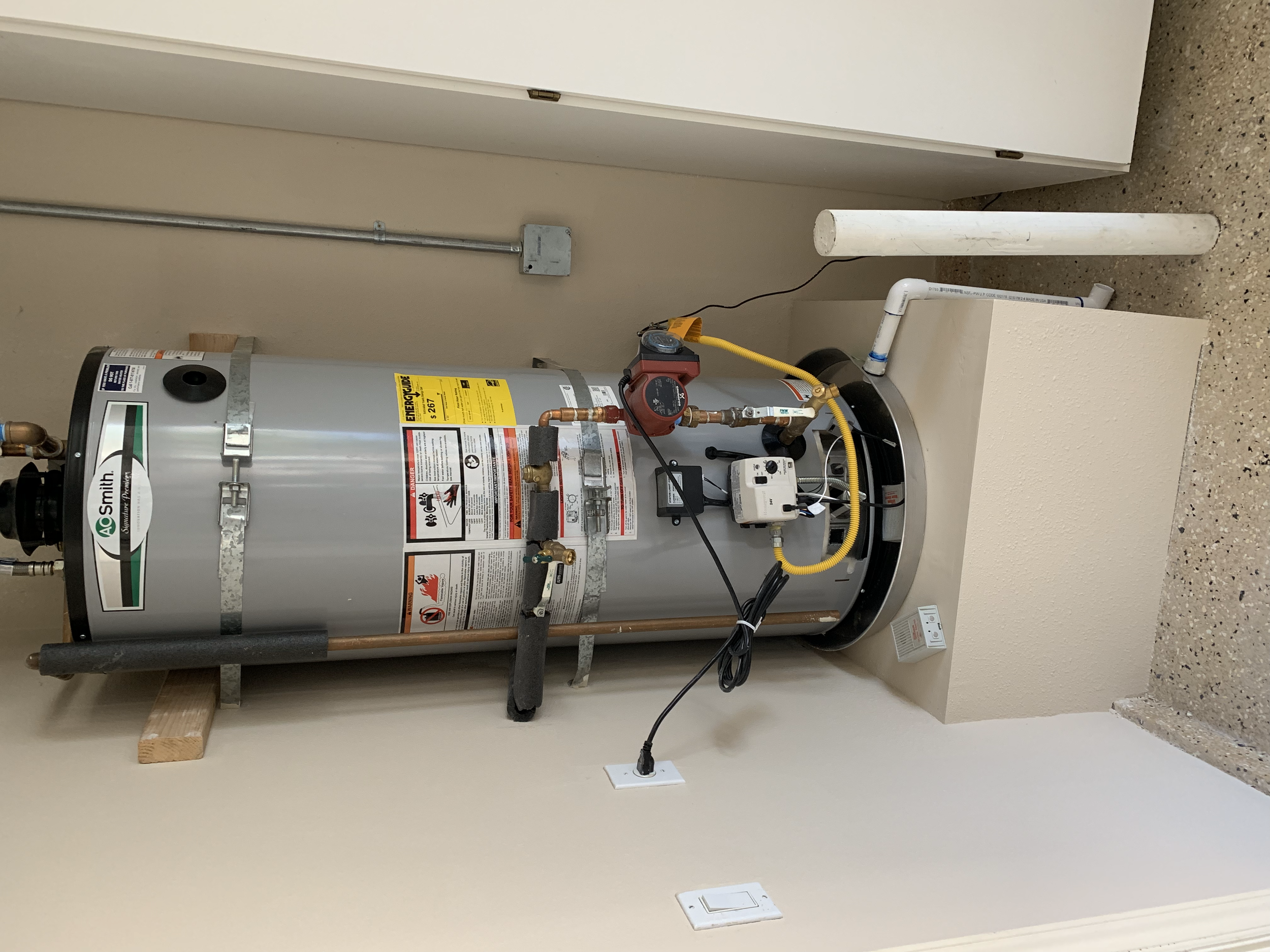 A water heater installed on a beige wall in a utility room, with pipes, electrical connections, and a red pressure gauge on top.