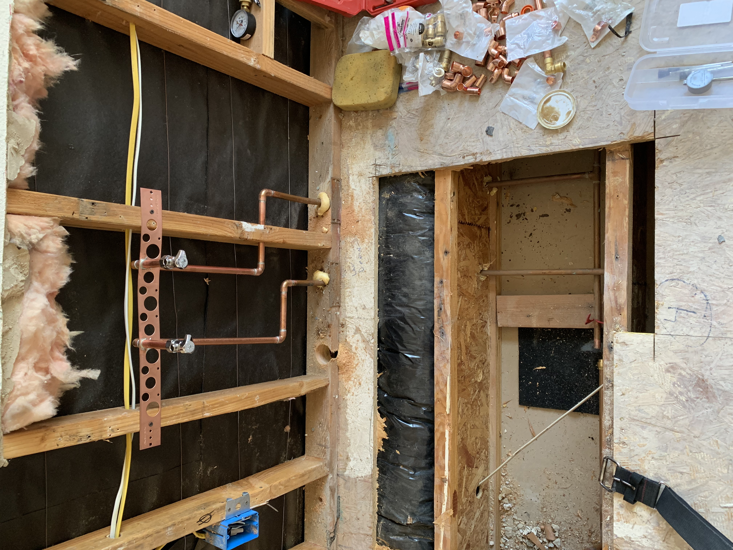 Partially constructed plumbing in a building with exposed wooden studs, copper pipes, and drainage pipes. Construction tools and materials are scattered above the opening in the floor, which is framed for a future fixture or utility installation.