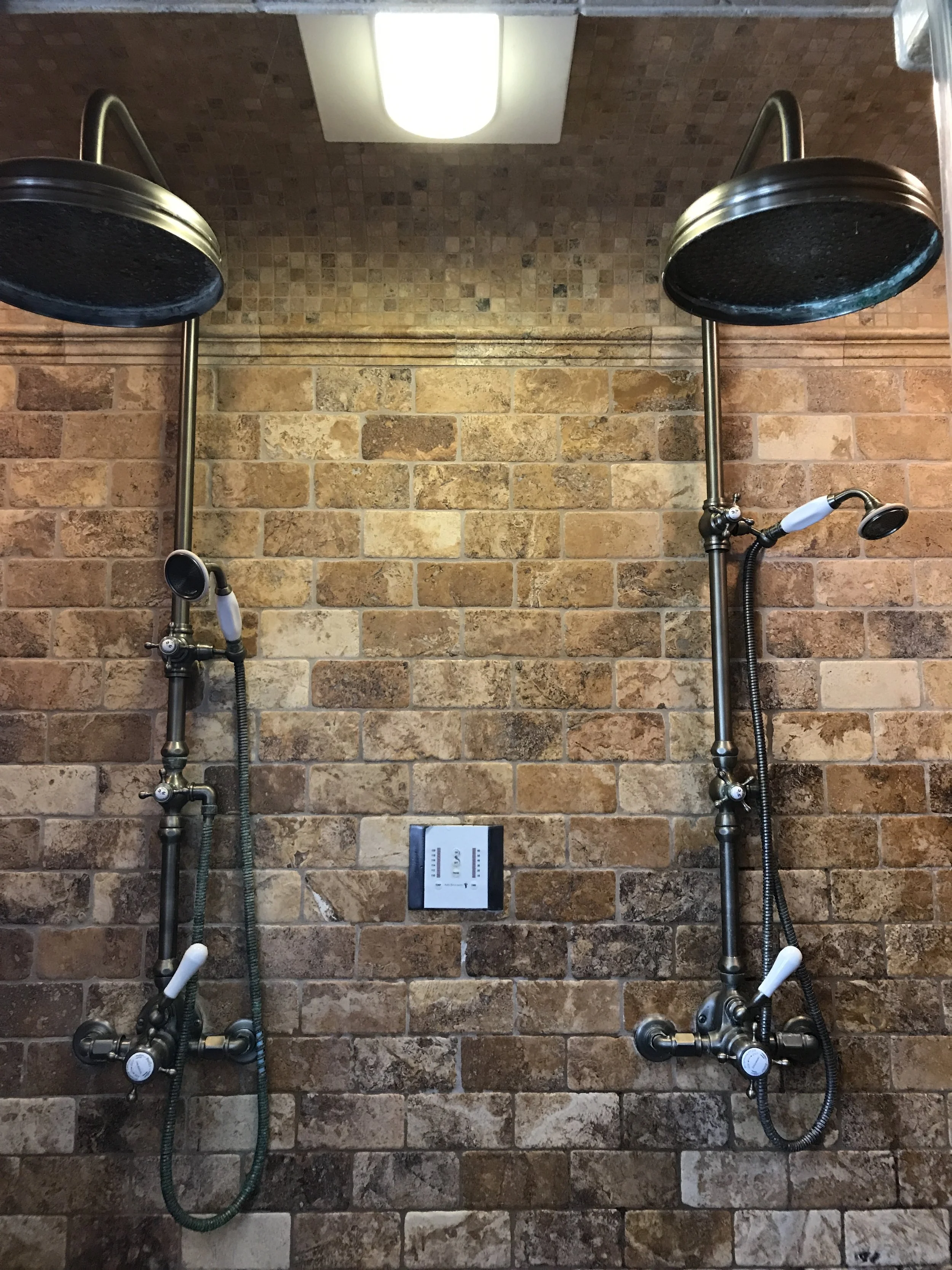 Two vintage-style showerheads with handheld spray attachments on a brick wall, with a white ceiling light above.