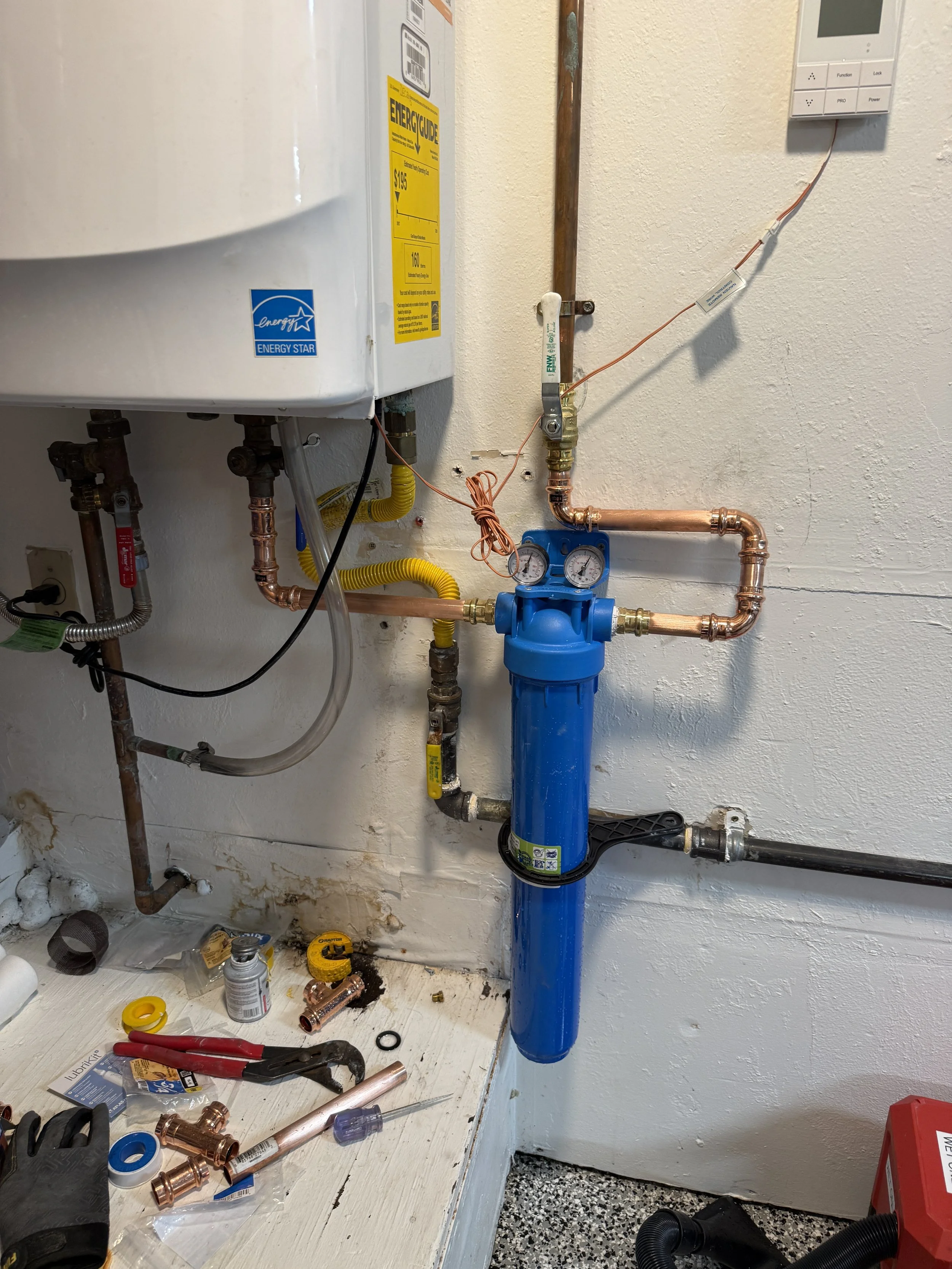 Tankless water heater sediment filter installation with blue filter in a utility room, copper pipes, gauges, tools, and miscellaneous plumbing supplies on a table.