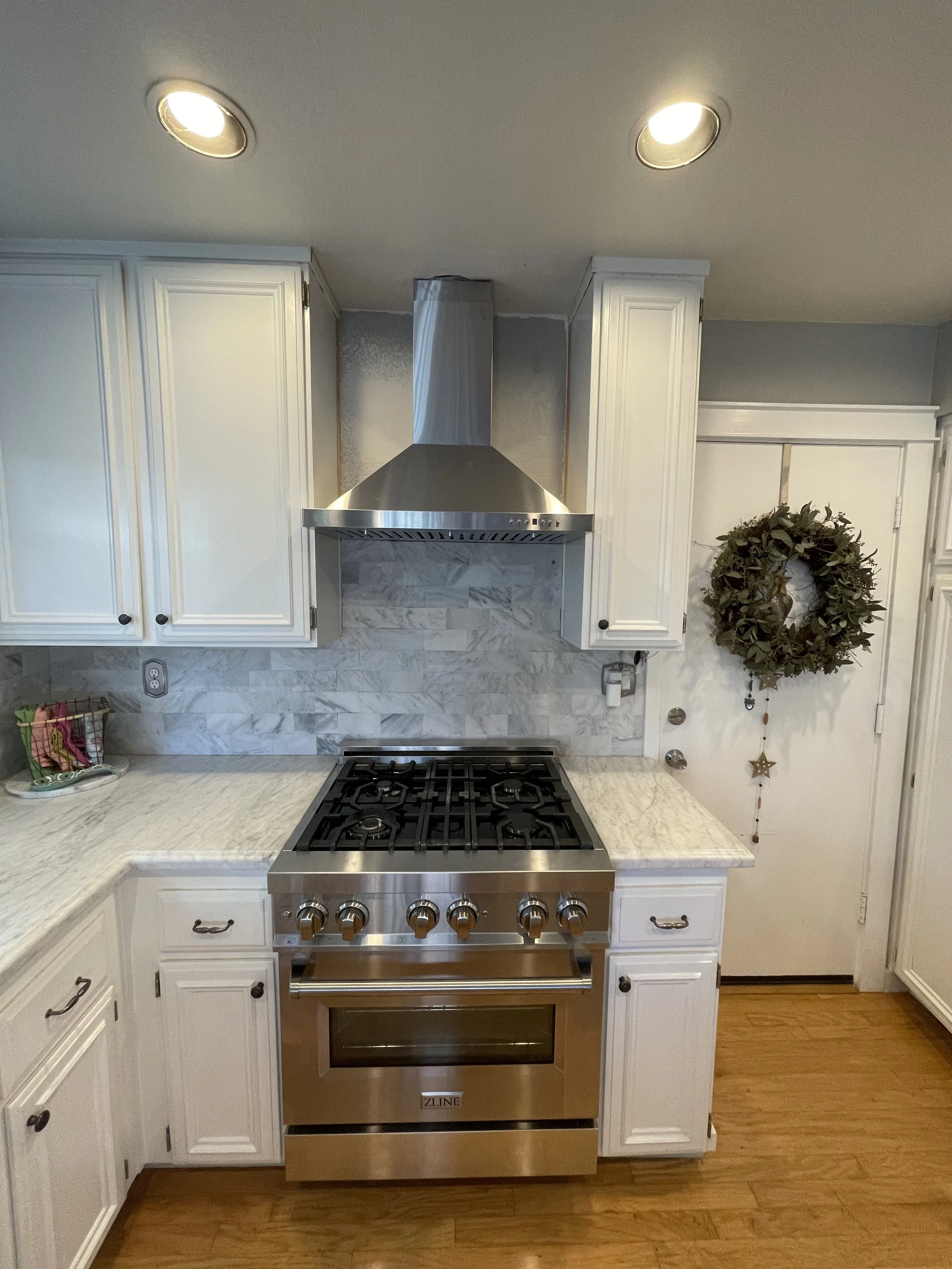 Kitchen with white cabinets, marble countertops, stainless steel stove and vent hood, backsplash, and wooden floor. Menorah wreath decor hangs on the white door on the right.