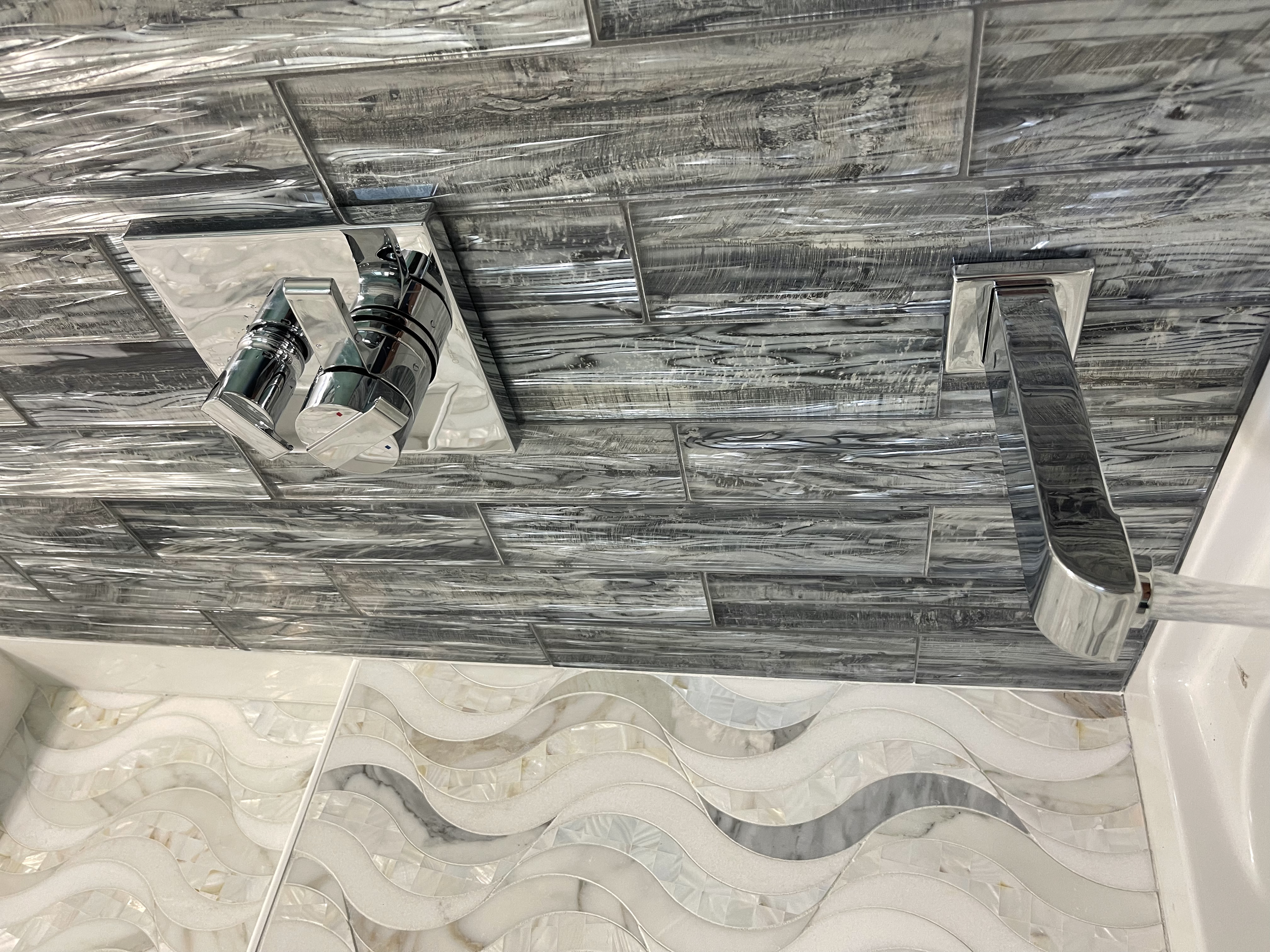 Close-up of a modern bathroom sink faucet with a sleek chrome finish, mounted on a wall with gray marble tiles and patterned beige and white tiles below.