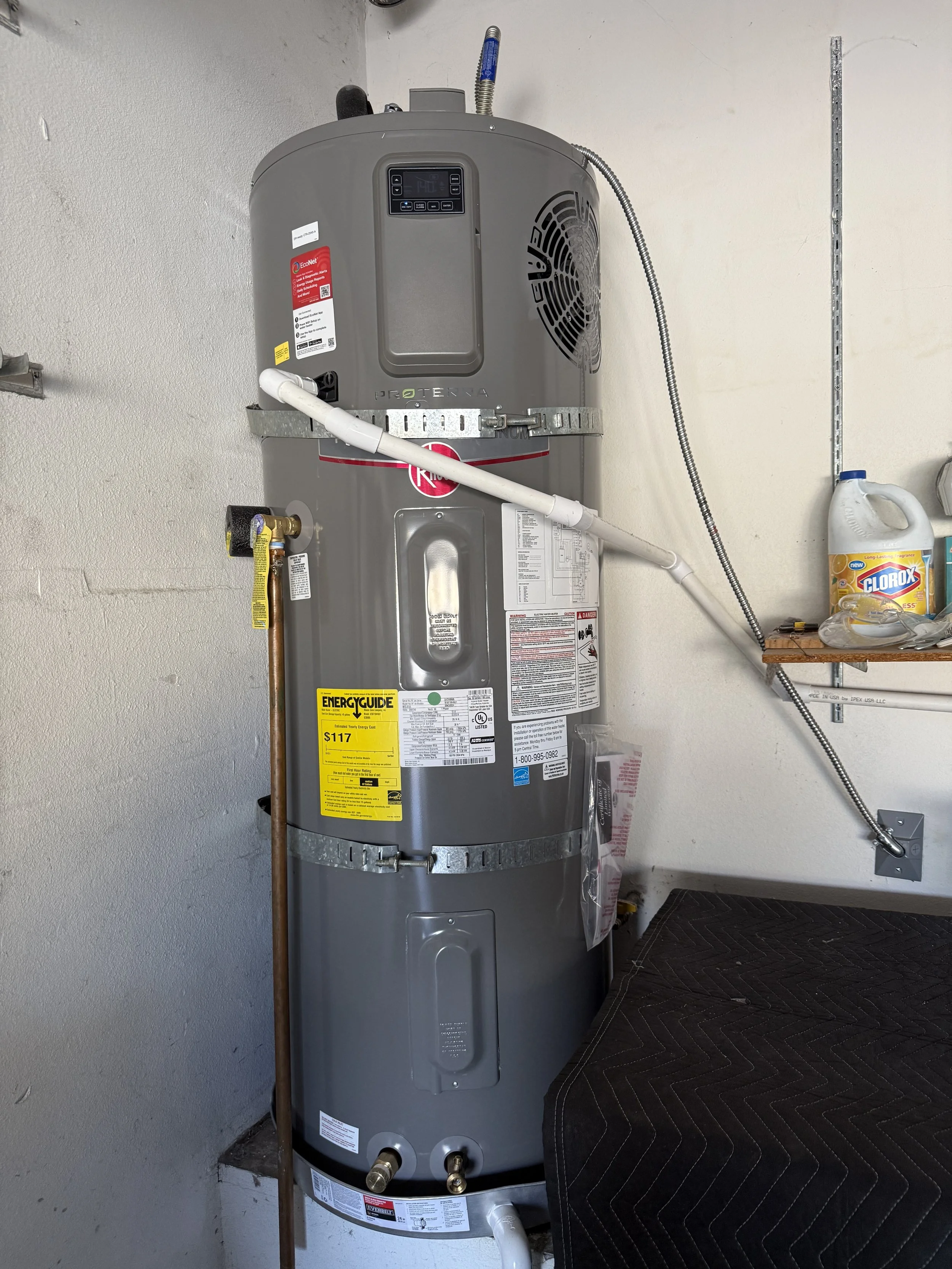 A tall, gray water heater in a garage or utility room, with pipes and labels attached, positioned next to a wall and a black cushioned surface.