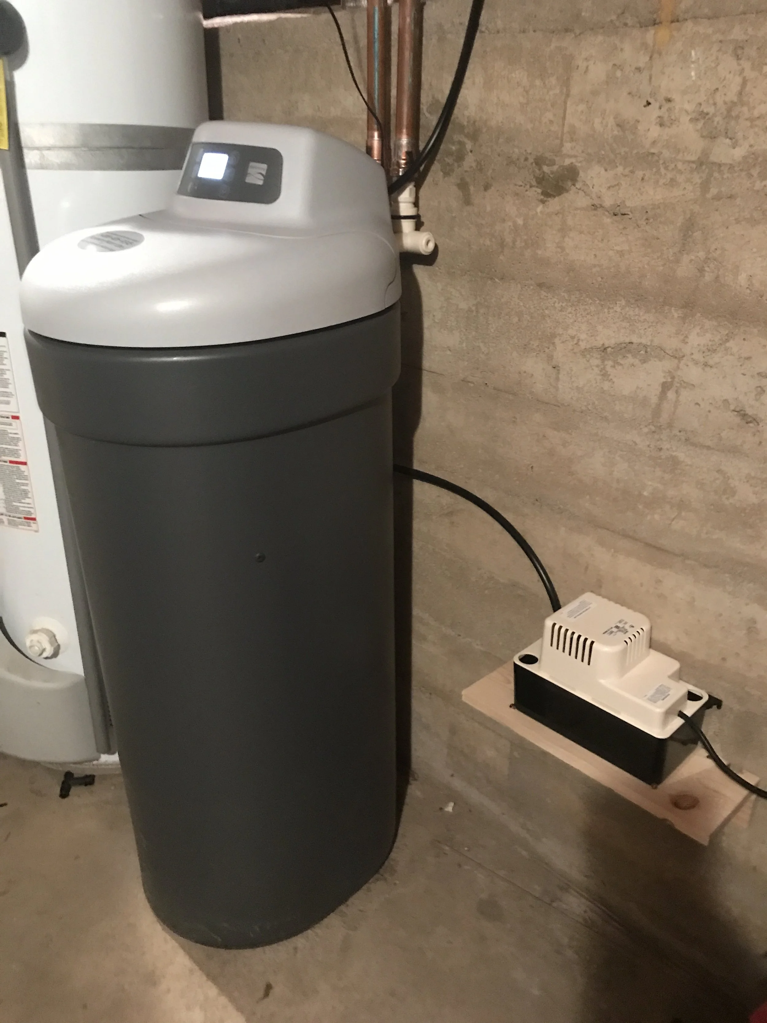 A water softener with a gray tank and a control panel on top, connected to pipes on a concrete wall, with a white electronic device installed on a small wooden shelf nearby.