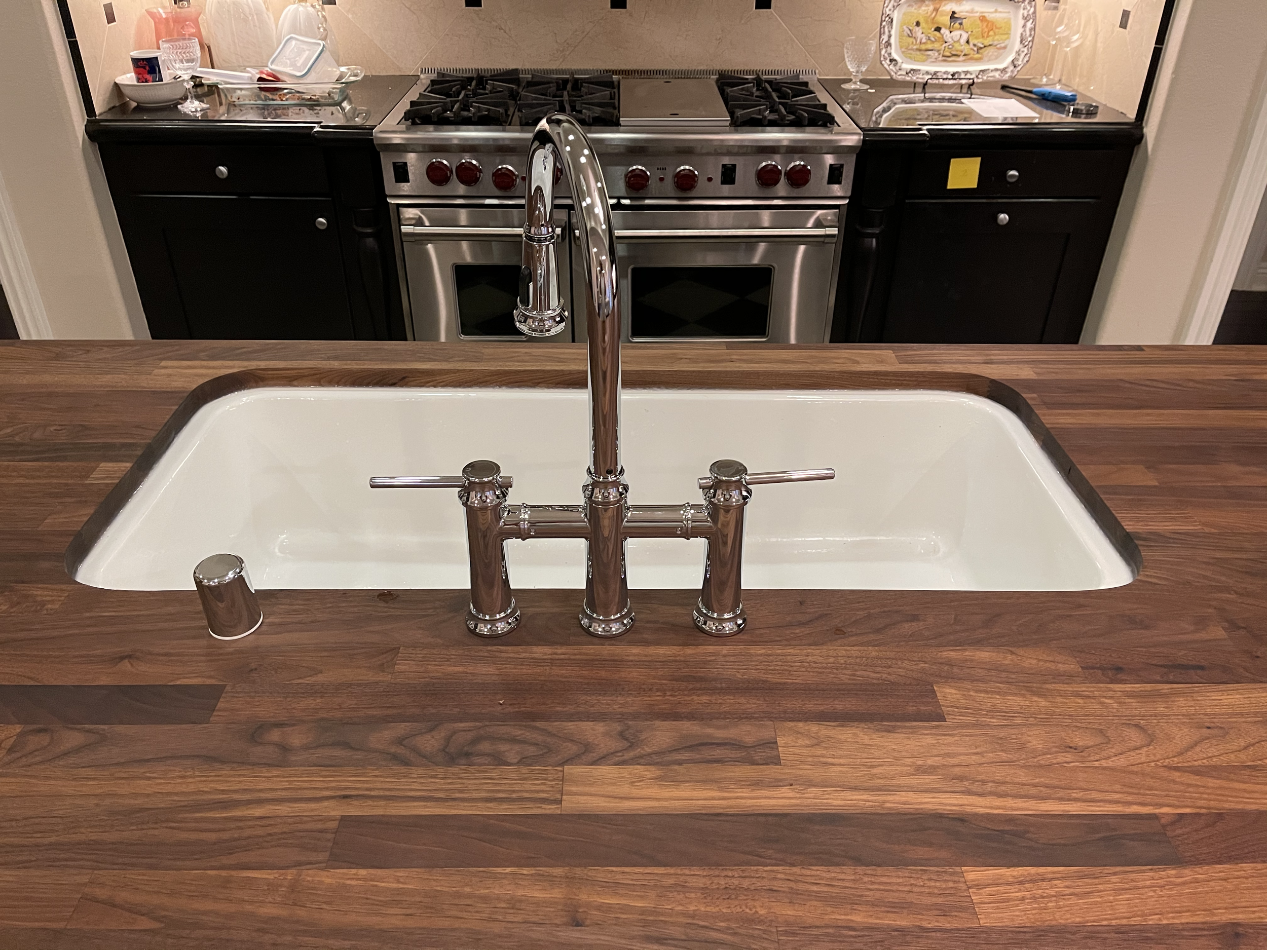 Undermount double porcelain cottage kitchen sink with a chrome faucet on a Walnut countertop, with a stove and black cabinets in the background.