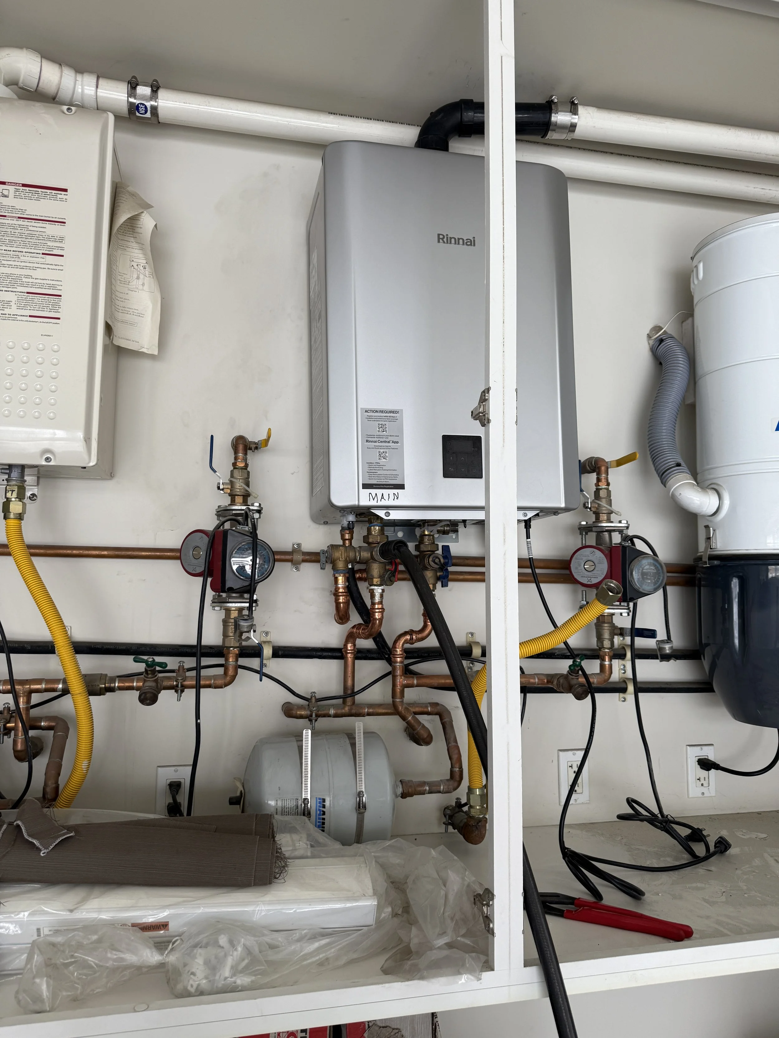 Indoor utility area with a Rinnai water heater, copper piping, gauges, electrical wires, and various tools and materials.