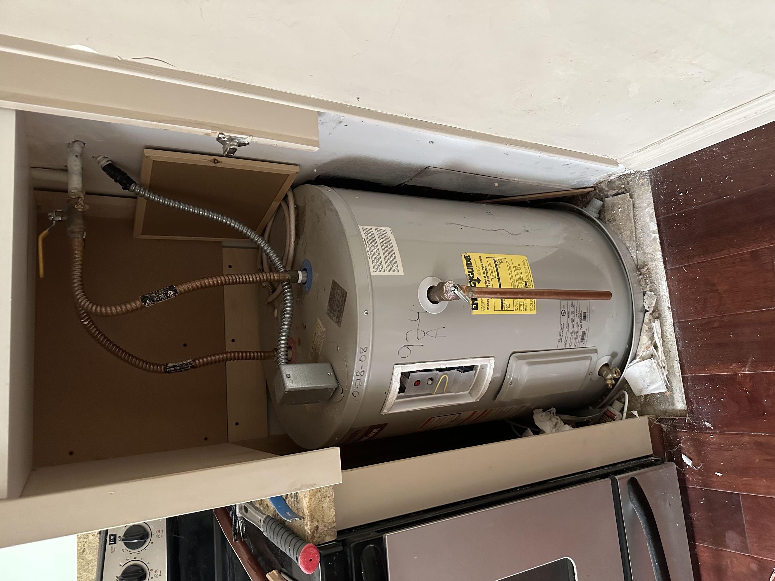 An electric water heater installed in a small cabinet area in a kitchen, with pipes and electrical connections visible.
