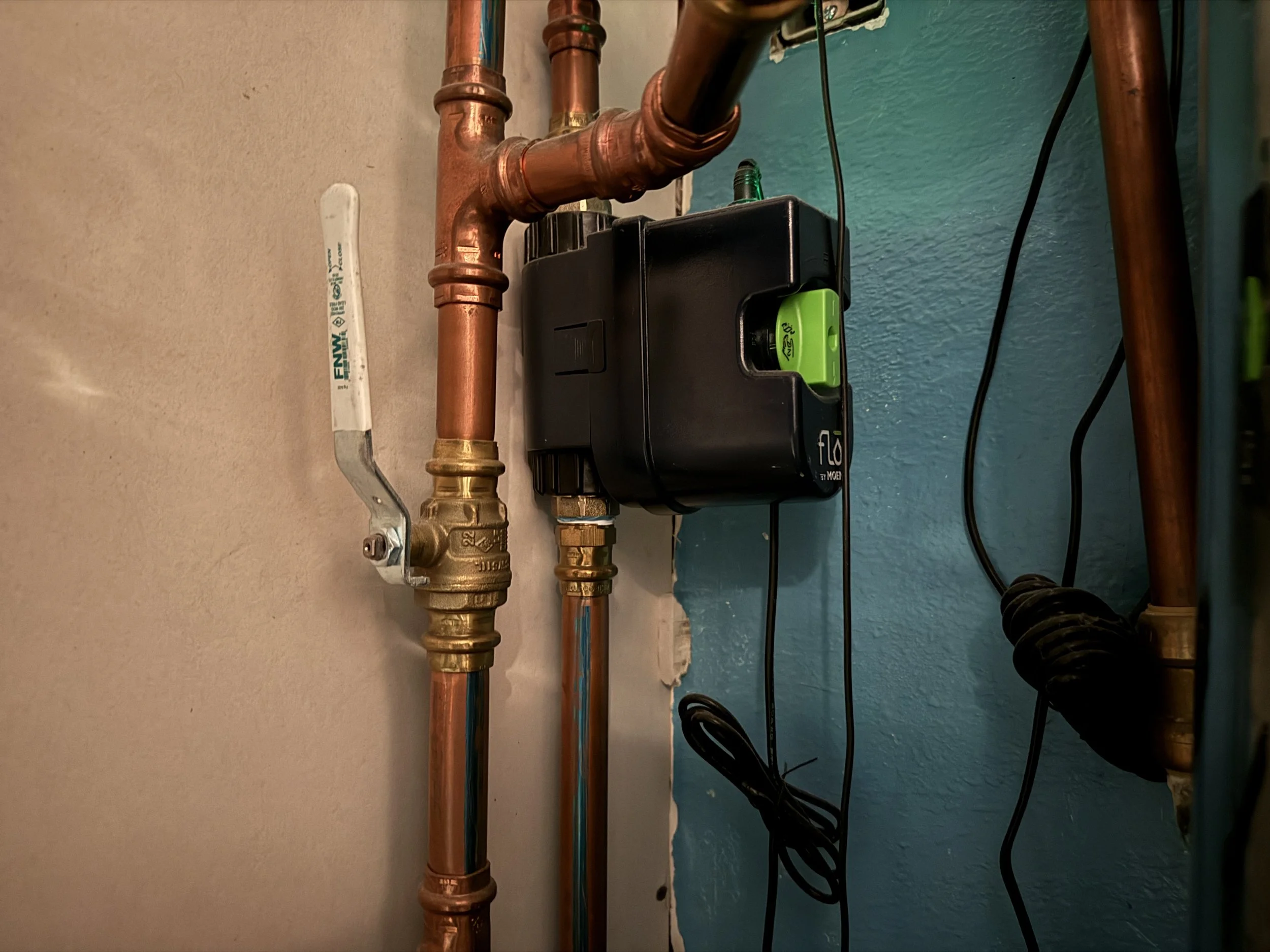 Copper Pipes with a Flo By Moen smart water shutoff valve system with a blue valve, connected to a black water pump with a green dial and a green switch, mounted on a wall with electrical wiring.