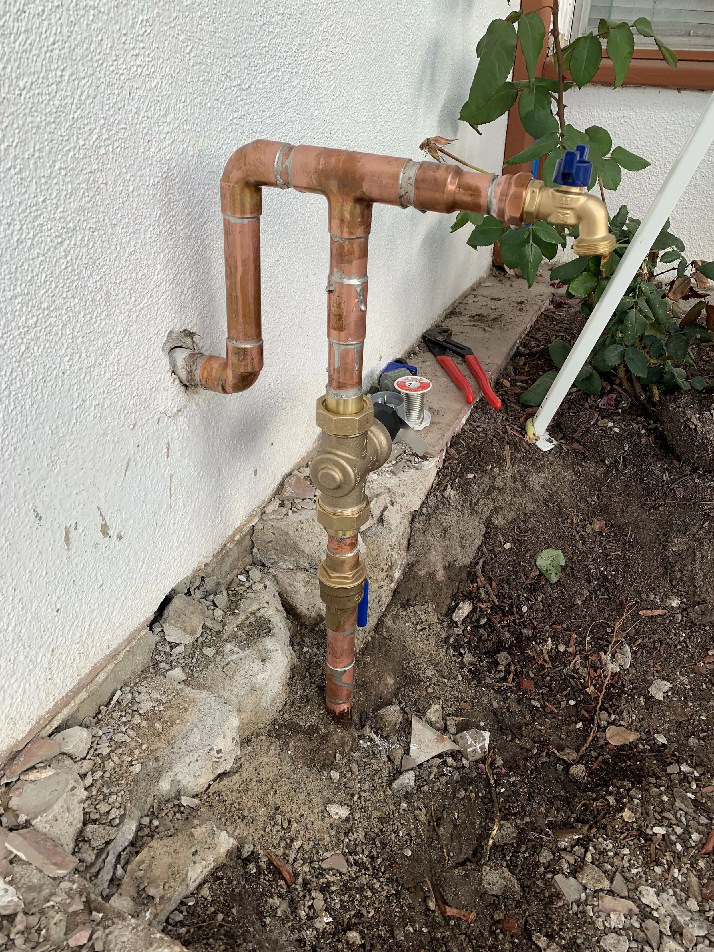 Plumbing service entry with copper pipes, a shut-off valve, and a faucet near a house wall, with gardening tools and soil nearby.