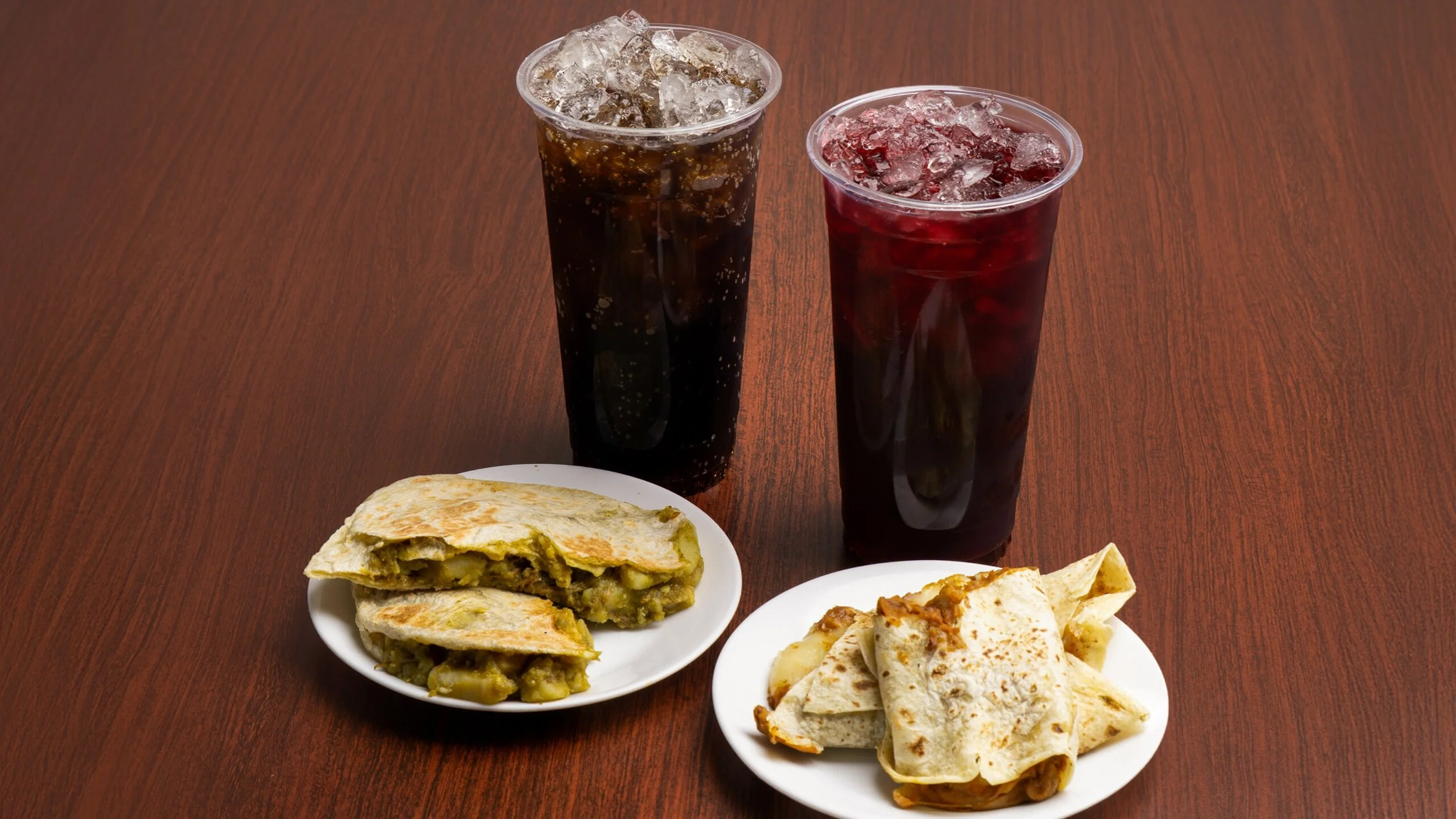 Two cups of soda with ice, one dark and one red, placed behind two plates of Mexican snacks on a wooden table. One plate has quesadillas with melted cheese and vegetables, and the other has folded tacos.