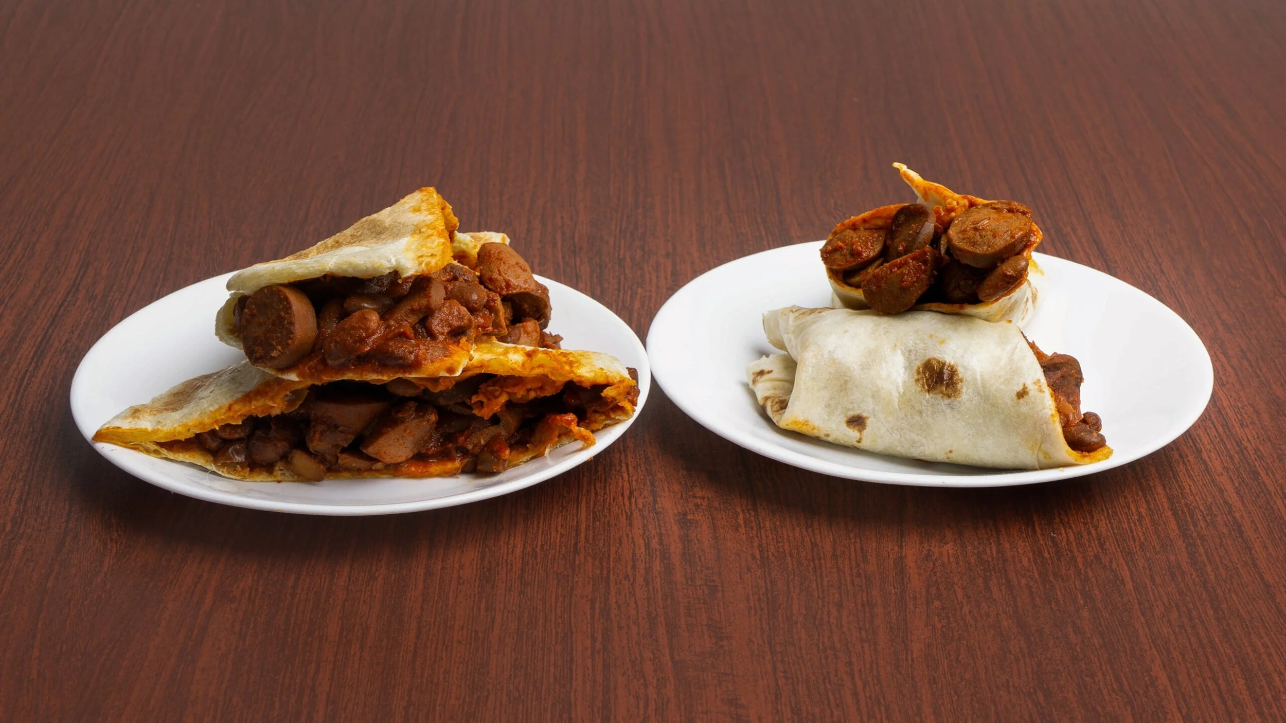 Two white plates containing bean burritos with chopped beans and sauce, on a dark wood table.