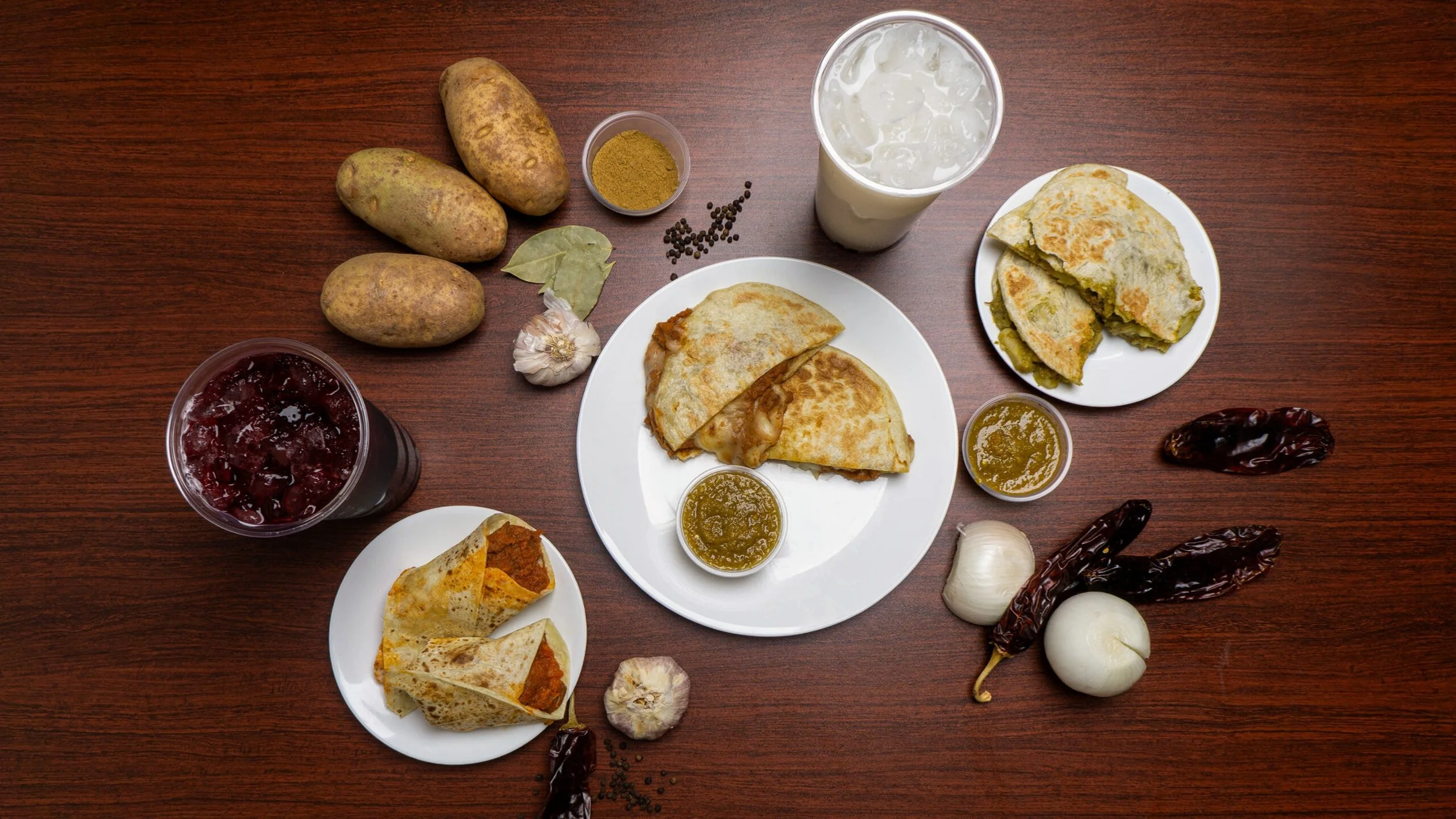 Indian meal featuring stuffed flatbreads served with green chutney, red chili peppers, garlic, and potatoes, accompanied by glass of iced drink and a glass of a cold white beverage.