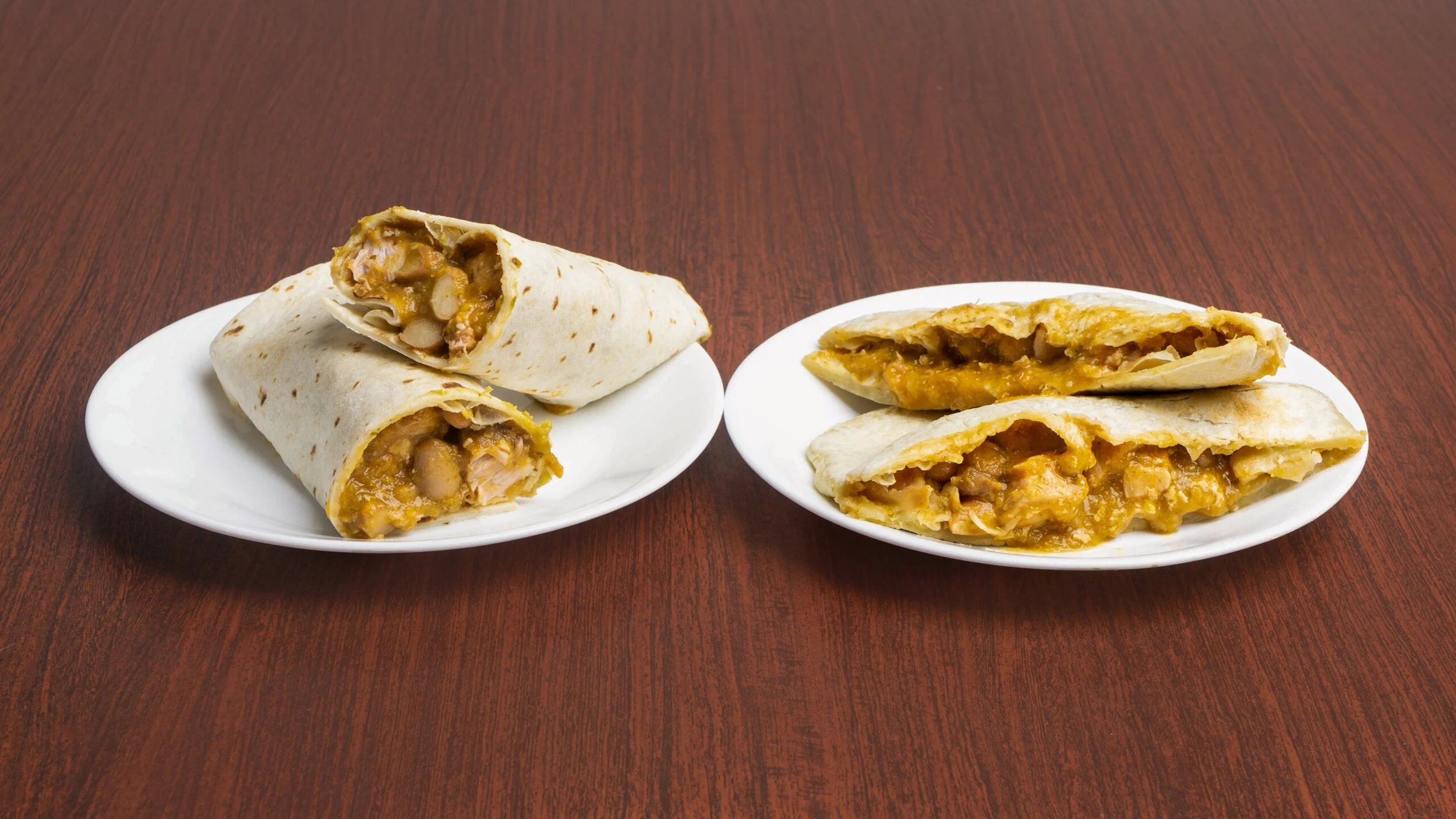 Two plates with burritos on a wooden table, one shows a whole burrito sliced in half revealing beans, chicken, cheese, and sauce; the other has two burrito halves cut open showing similar filling.